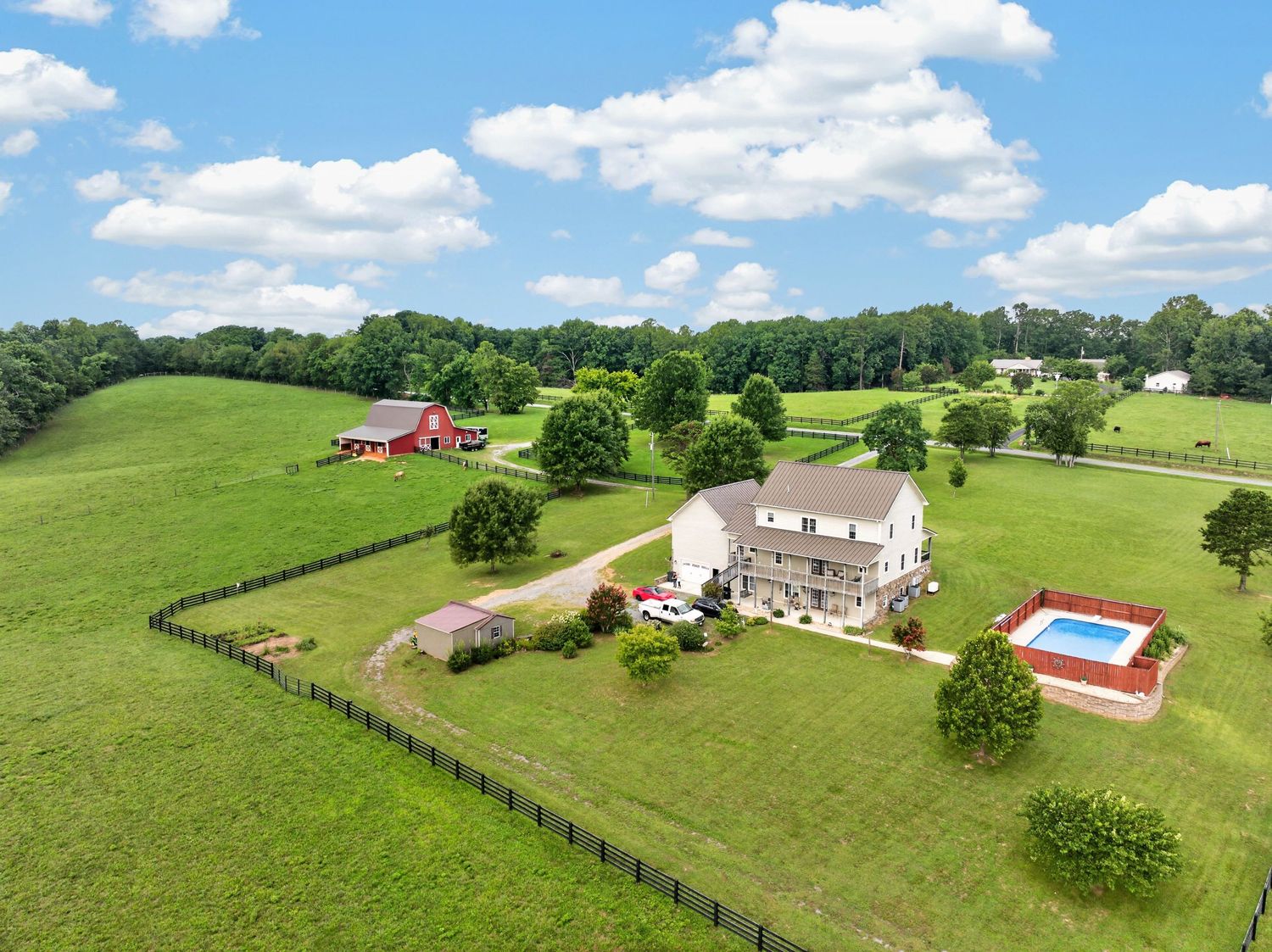 Aerial view of a rural farm property with a white house, red barn, pool, and lush green pastures surrounded by fencing.