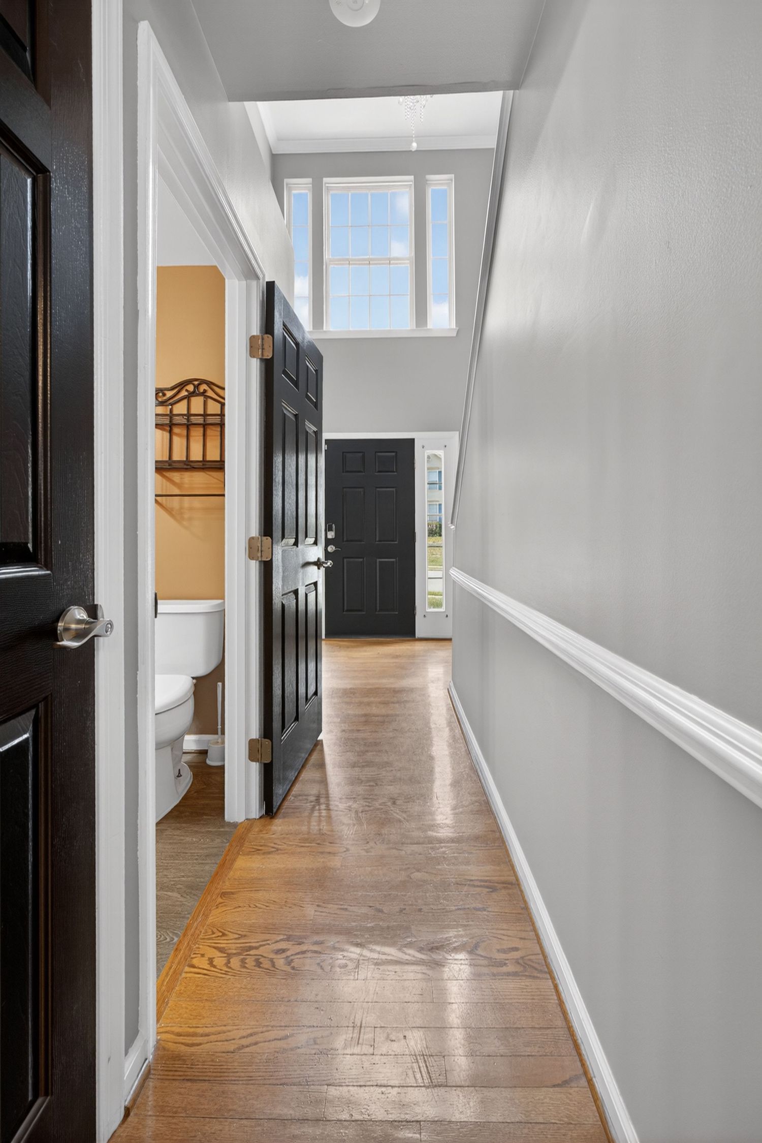 Bright hallway with hardwood floors, gray walls, black doors, and natural light from a transom window leading to the front door.