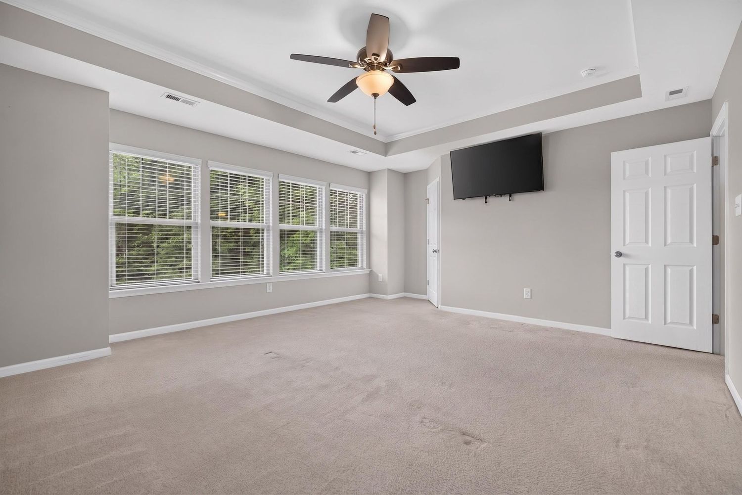 Spacious bedroom with tray ceiling, ceiling fan, wall-mounted TV, large windows, and neutral carpet flooring.