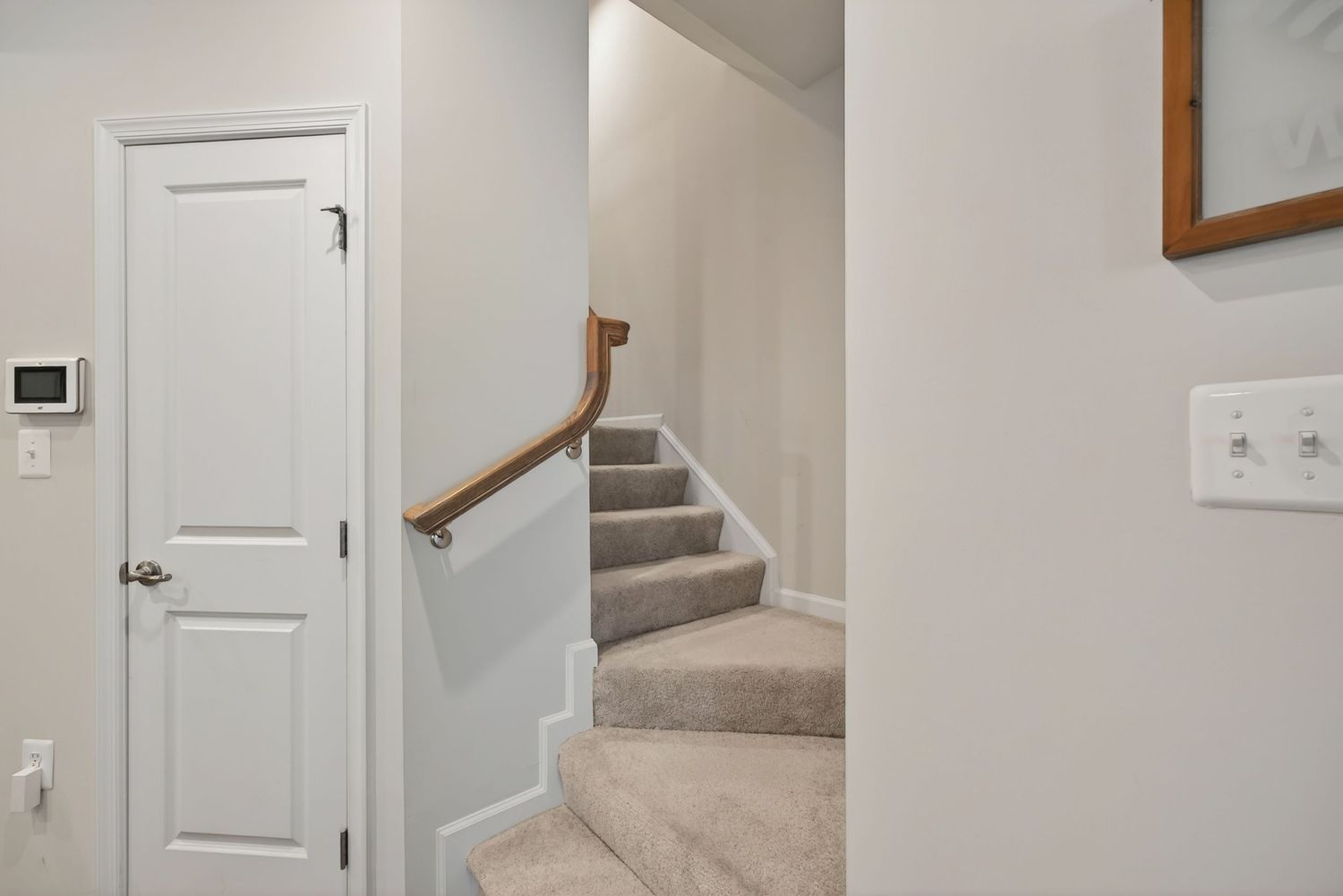 Bright hallway with white door, carpeted staircase, wood railing, and neutral walls in a modern home interior.