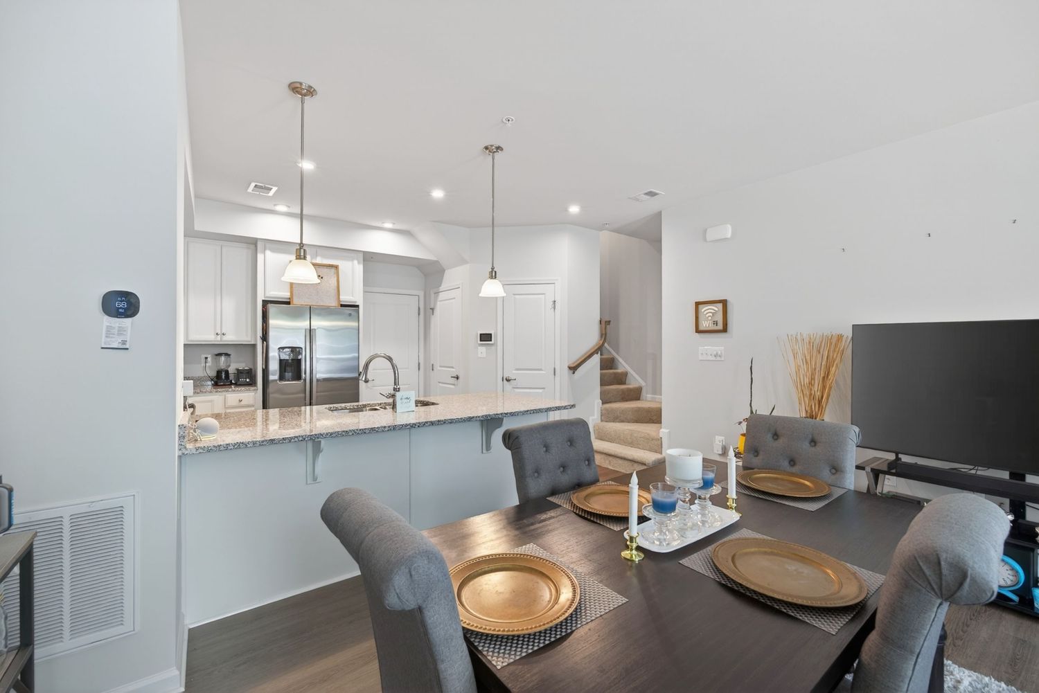 Modern open-concept dining area with gray chairs, gold placemats, and a white kitchen featuring stainless steel appliances.