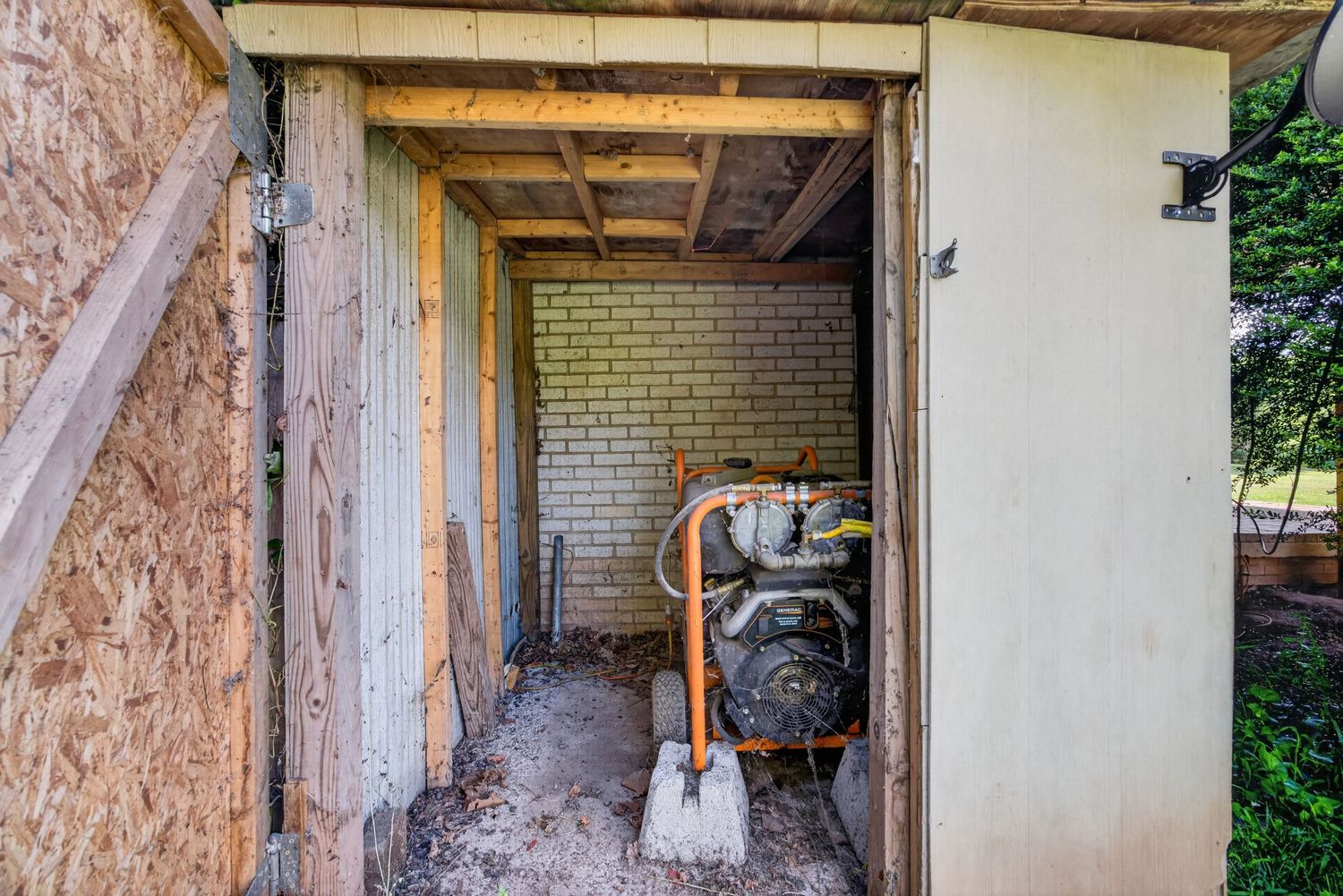 Small outdoor utility shed with brick wall, exposed wooden beams, and mechanical equipment including a pump on dirt floor.