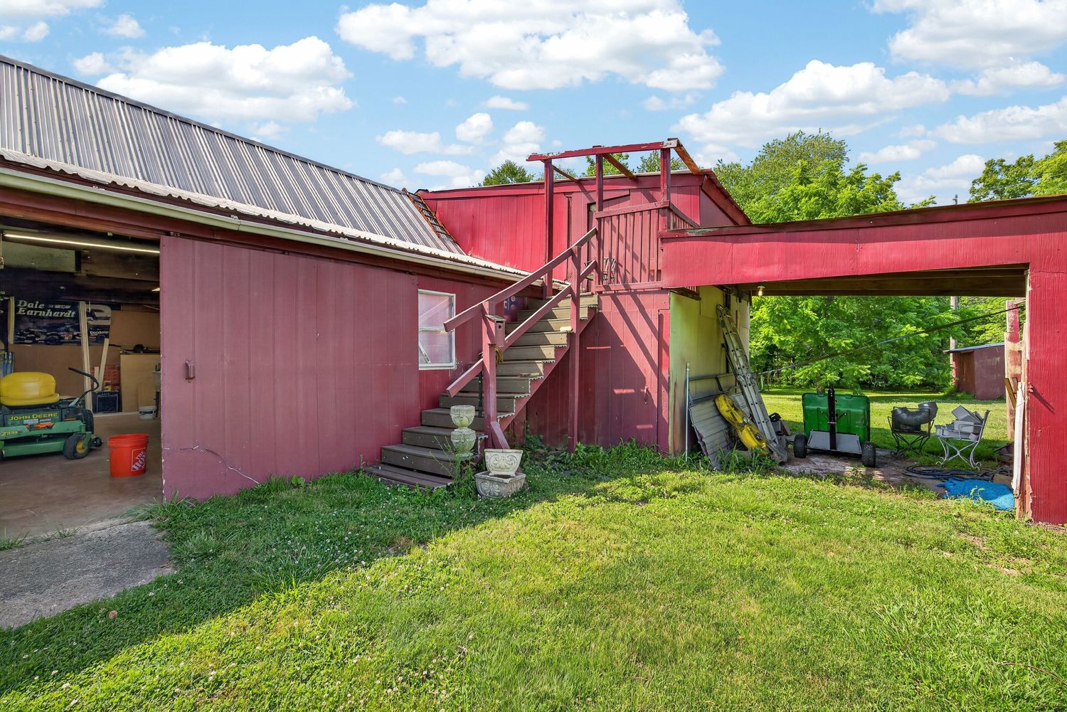 Red barn-style building with metal roof, exterior staircase, carport, and lush green lawn on a sunny day.