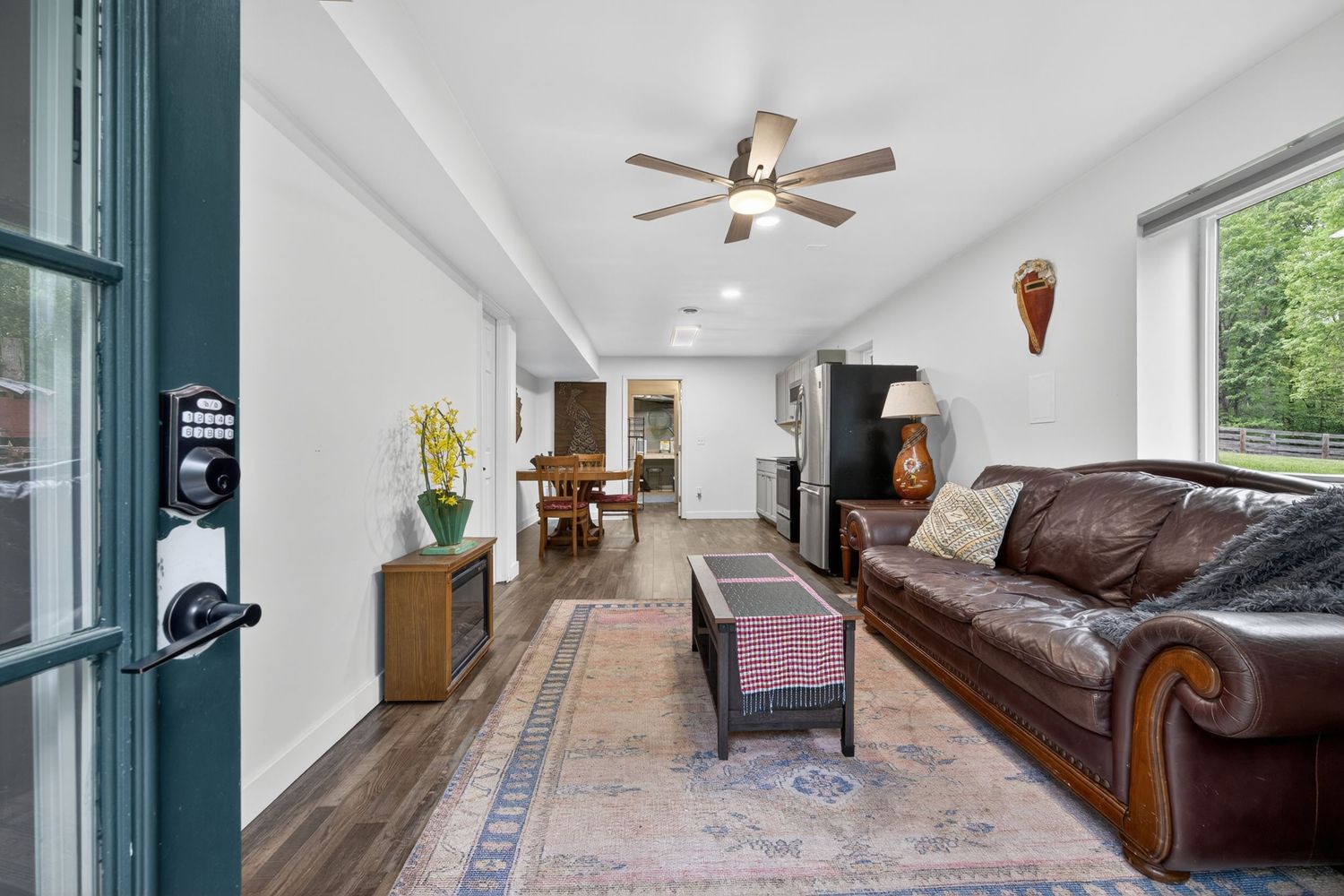 Bright living room with leather sofa, ceiling fan, hardwood floors, and area rug leading to dining area with natural light.