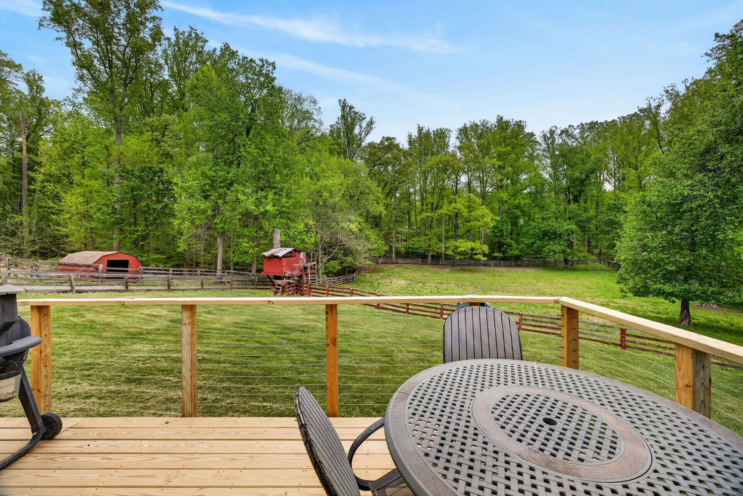 Wooden deck with patio table and chairs overlooking a lush green yard with red barn and tall trees.