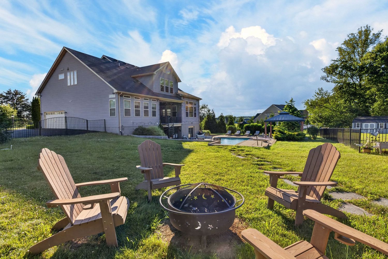 Backyard with Adirondack chairs around a fire pit, lush green lawn, pool, and large house under a blue sky.