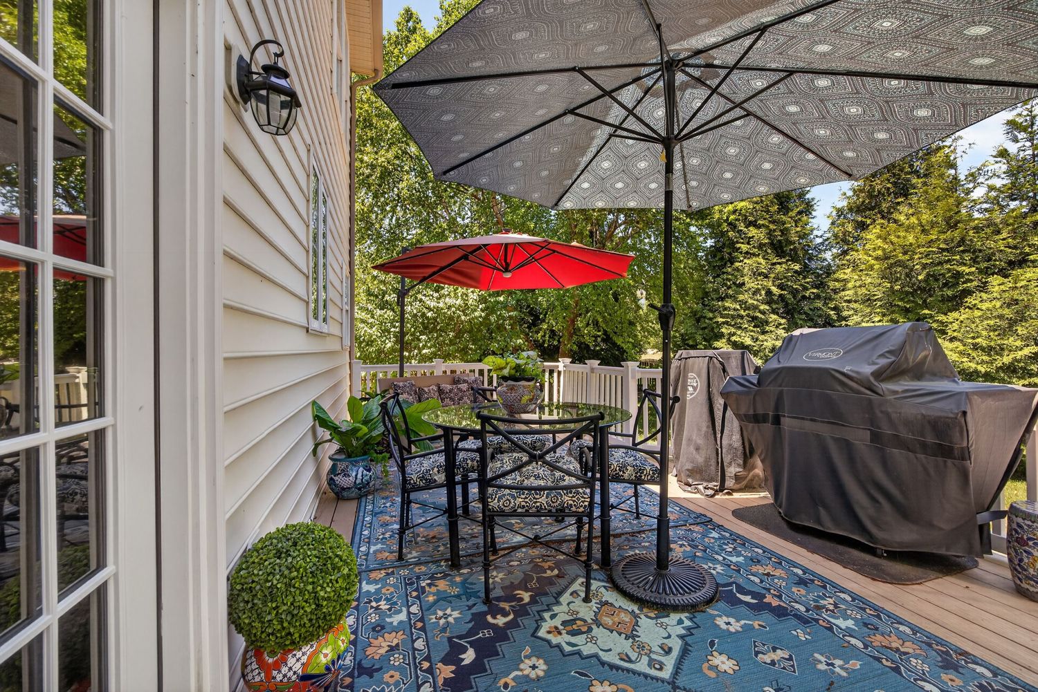 Outdoor deck with patio table, red umbrella, blue floral rug, and gas grill surrounded by lush green trees.