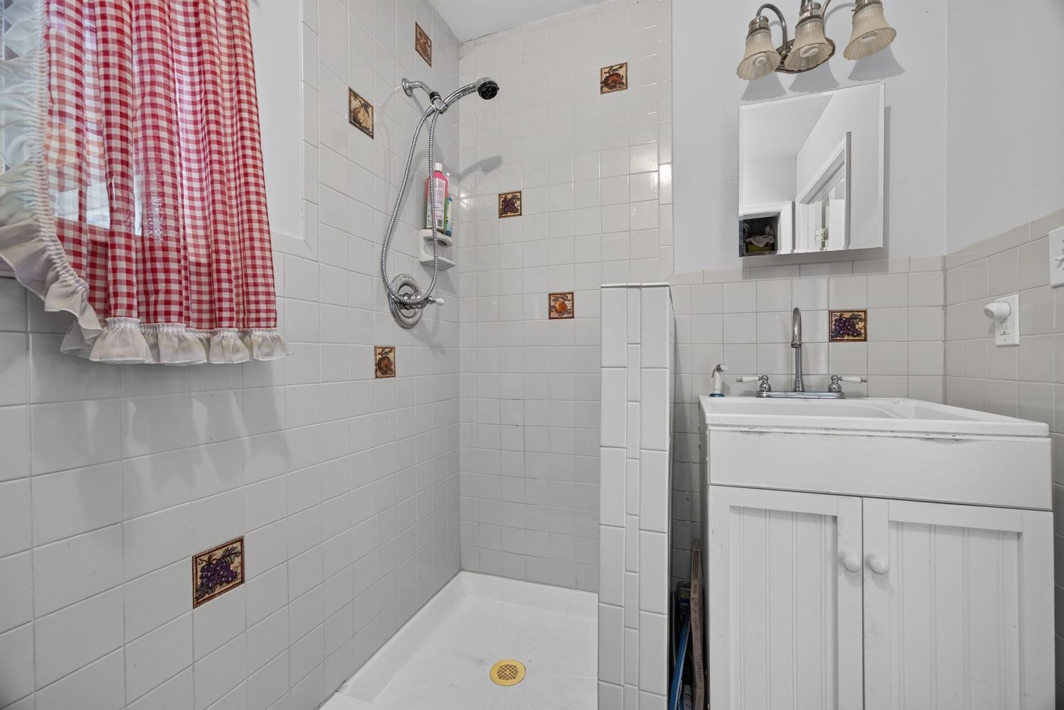 White tiled bathroom with walk-in shower featuring red gingham curtain, decorative accent tiles, and white vanity with medicine cabinet.