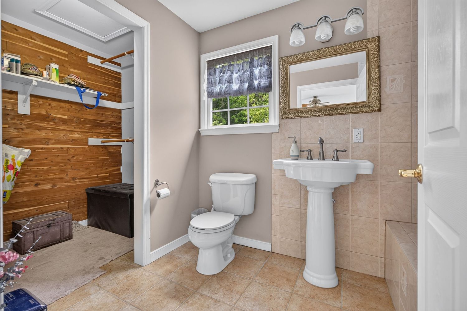 Bright bathroom with pedestal sink, toilet, framed mirror, and wood-paneled walk-in closet on tile flooring.