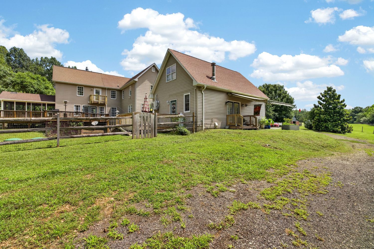 Rural home with large green lawn, wooden fence, and deck surrounded by lush trees under a blue sky.
