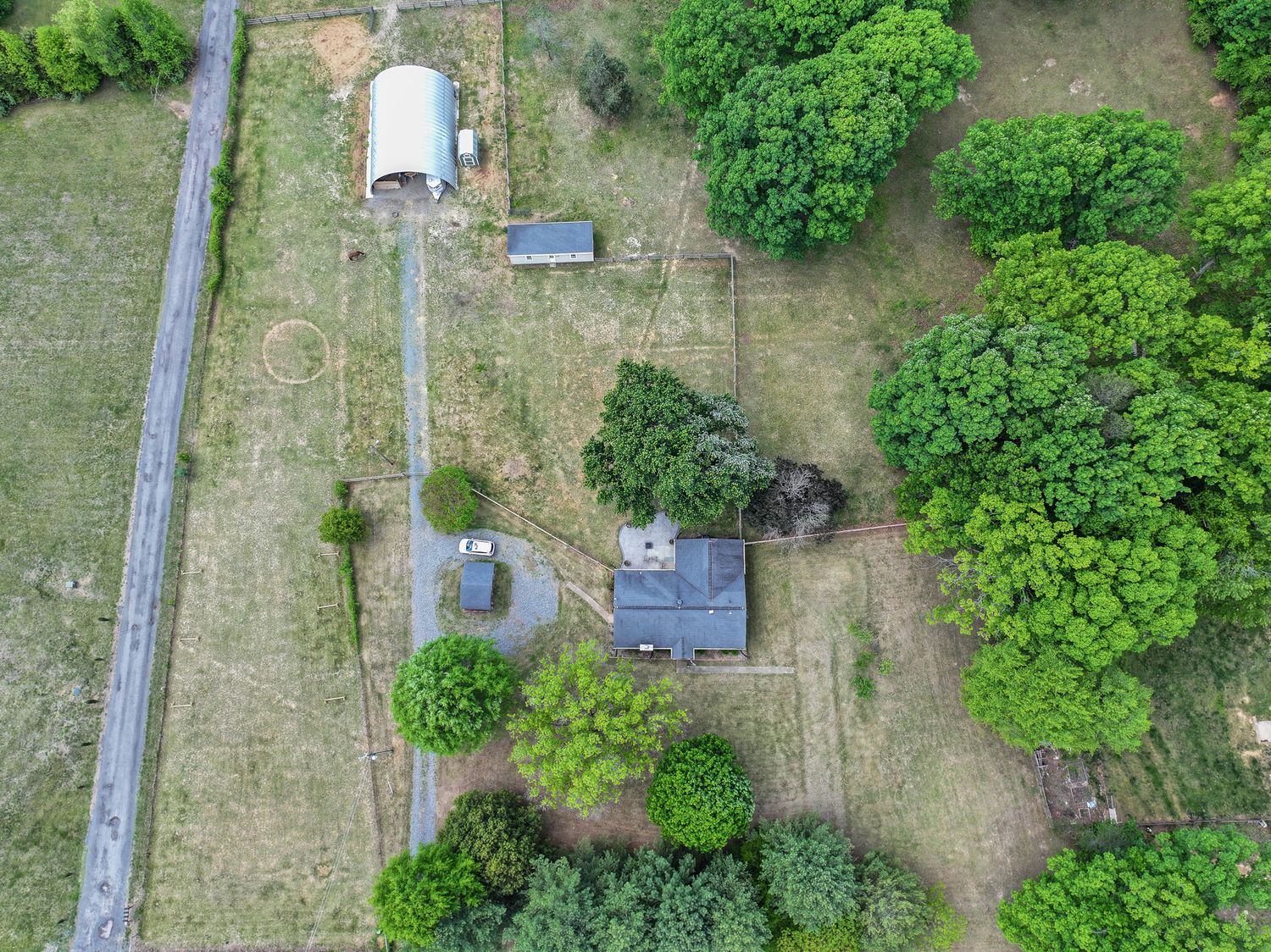 Aerial drone view of a rural property featuring a metal barn, small house, and lush green trees surrounded by open farmland.