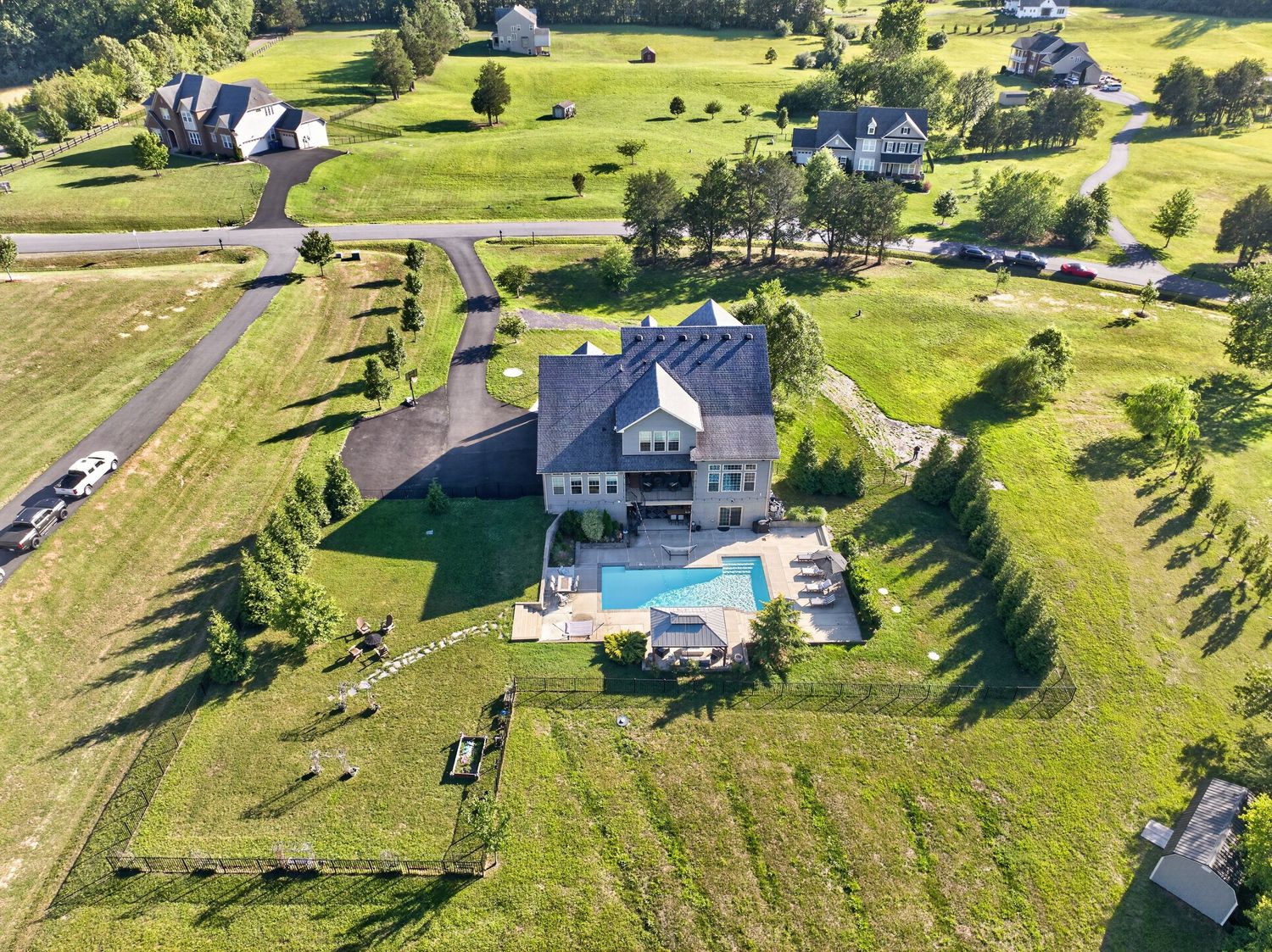 Aerial view of a large luxury home with a pool surrounded by lush green fields and trees in a rural neighborhood.