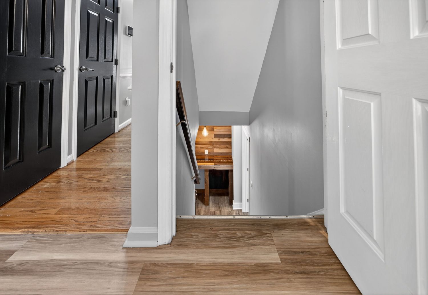 Bright hallway with hardwood floors, white walls, black door, and staircase leading to lower level.
