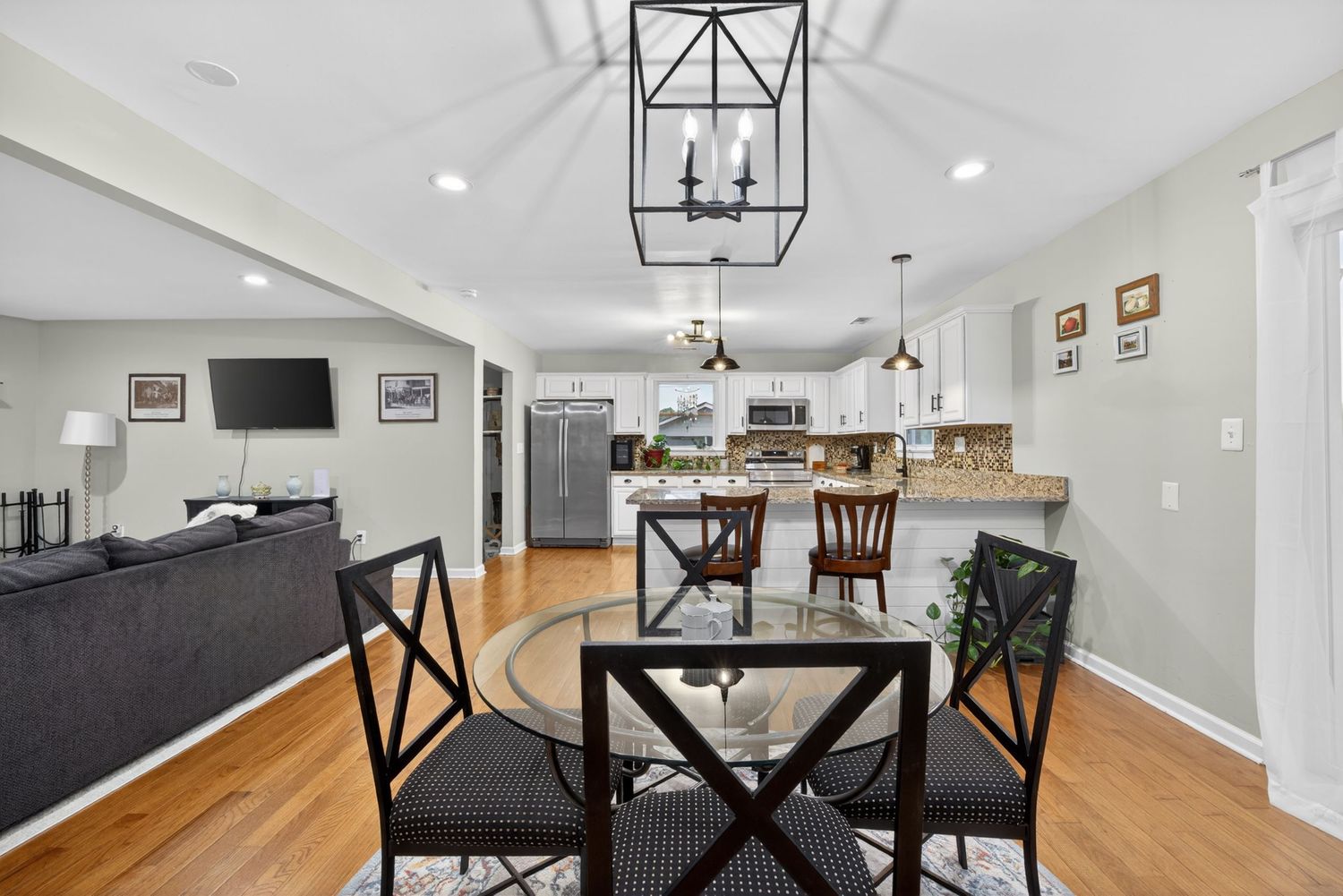 Open-concept dining area with glass table, black chairs, hardwood floors, and a modern chandelier leading to a white kitchen.