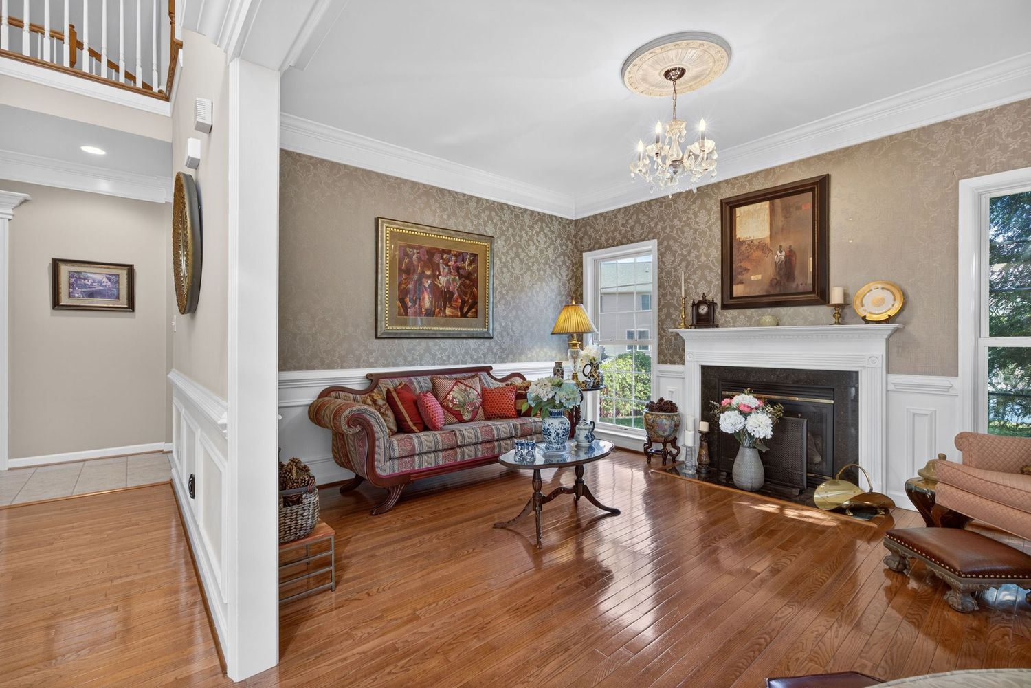 Elegant living room with hardwood floors, ornate fireplace, chandelier, floral wallpaper, and classic furniture in a traditional home.