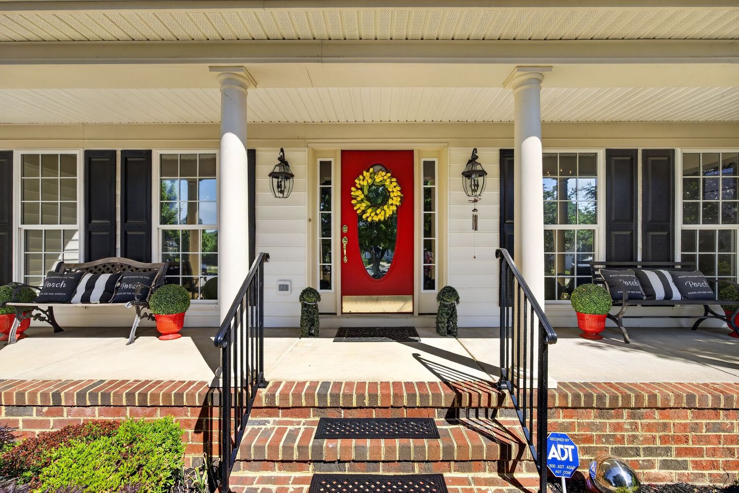 Charming farmhouse front porch with red door, wreath, white columns, brick steps, and black railings.