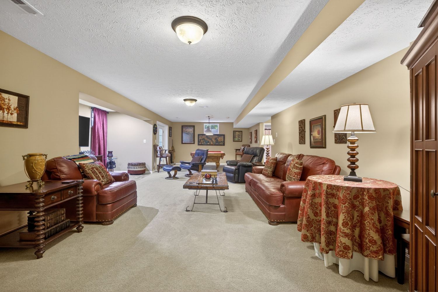 Spacious basement living room with leather sofas, carpet flooring, and warm neutral walls under a textured ceiling.