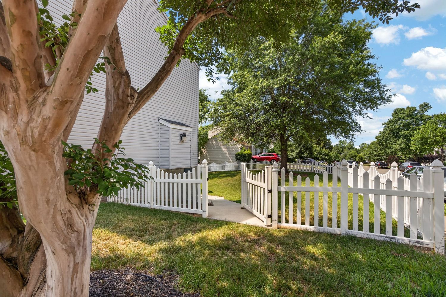 White picket fence gate leading to a lush green yard with mature trees beside a white suburban home.