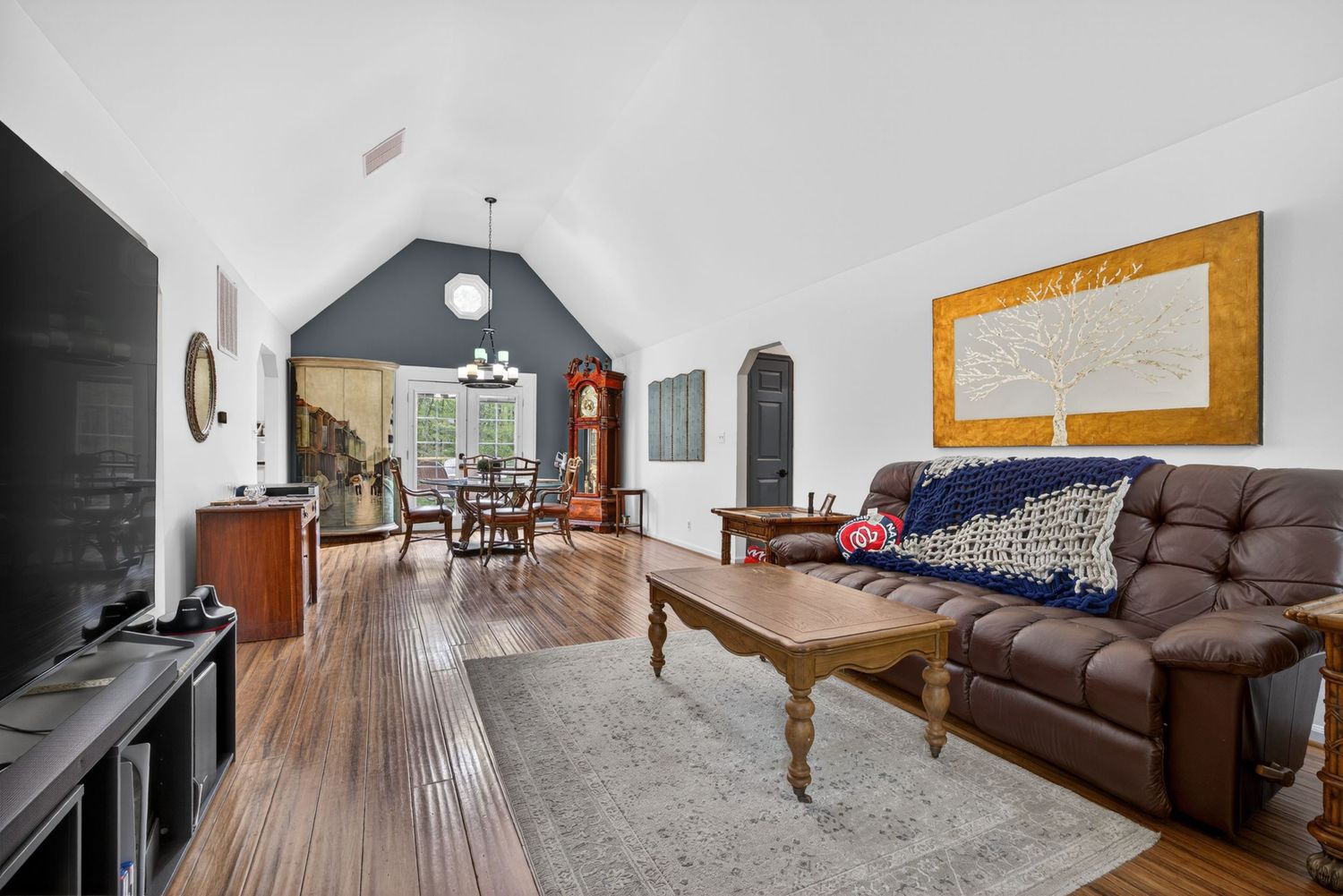 Spacious living room with vaulted ceilings, hardwood floors, leather sofa, wood coffee table, and dining area in background.