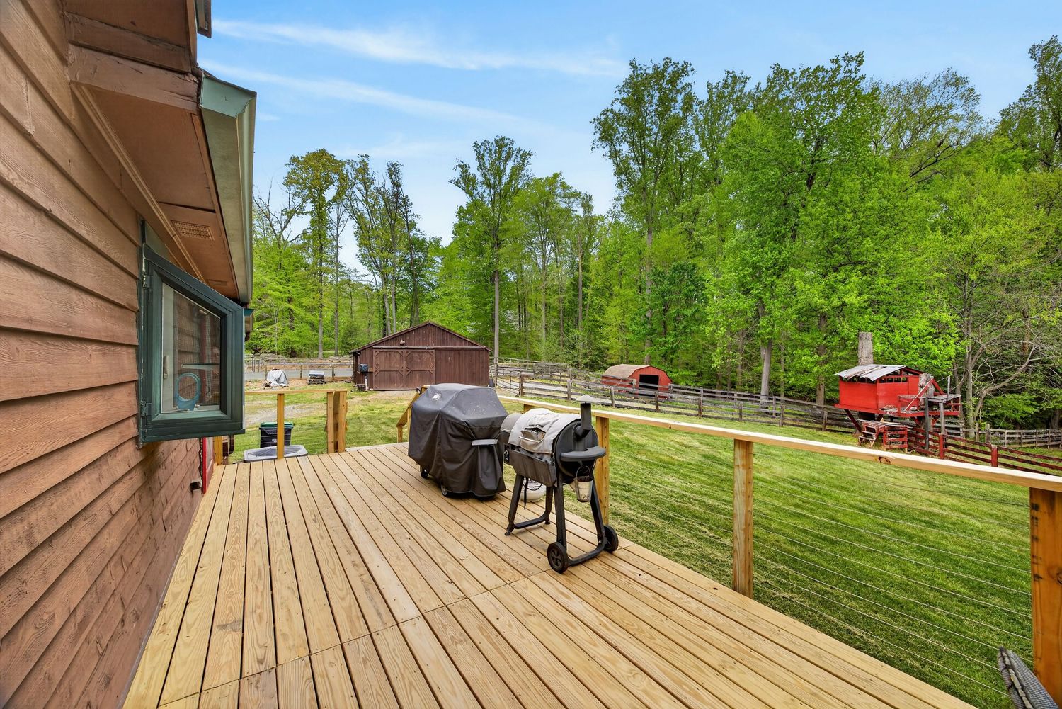 Wooden deck with a grill overlooking a lush green backyard with a barn and red structure surrounded by trees.