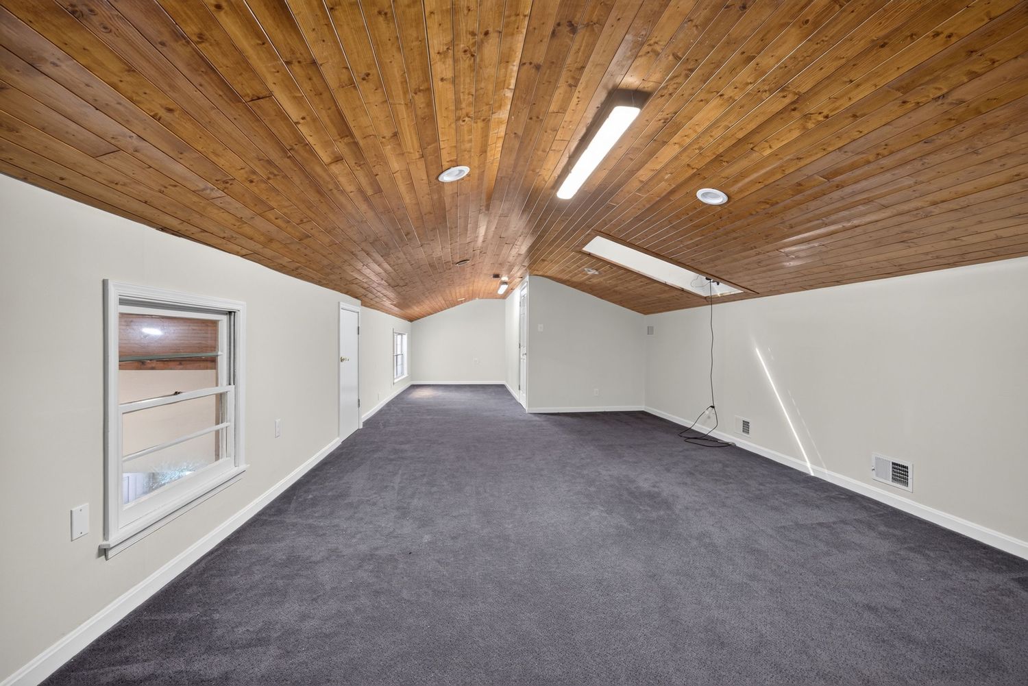 Spacious empty room with vaulted wood ceiling, skylights, dark carpet, and white walls with a pass-through window.