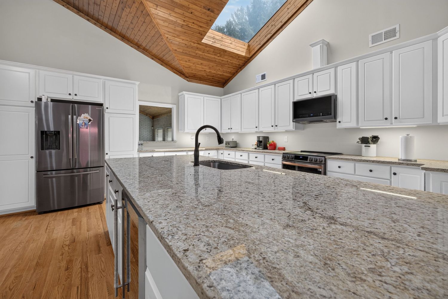 Modern kitchen with granite island, stainless appliances, white cabinets, and wood ceiling.