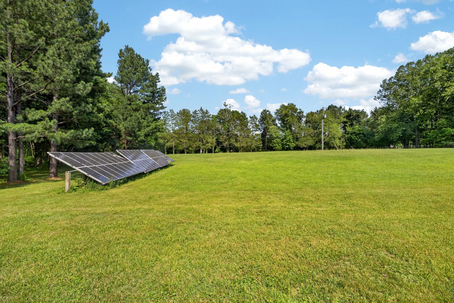 Ground-mounted solar panels on a lush green lawn surrounded by tall trees under a partly cloudy blue sky.