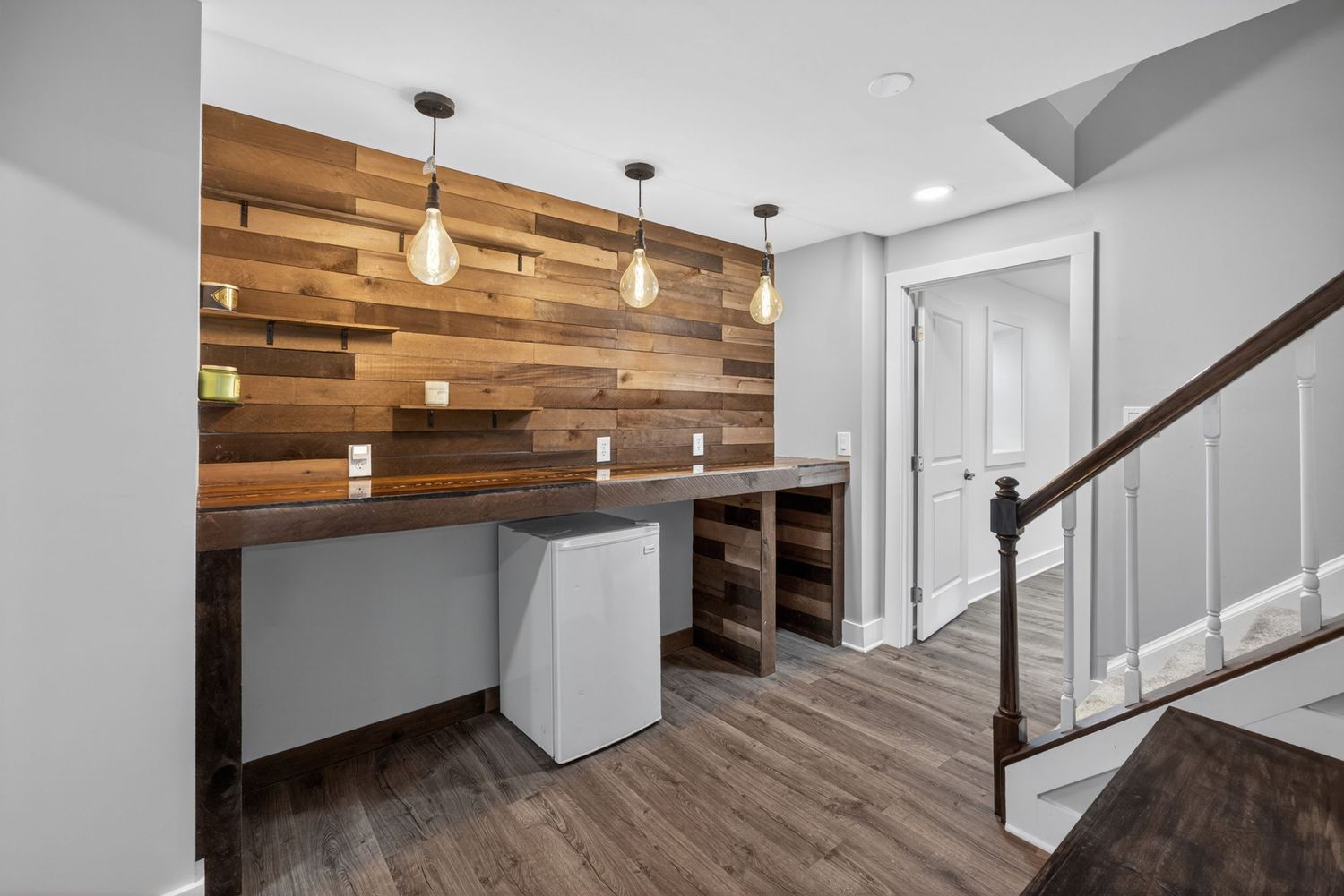 Modern basement wet bar with reclaimed wood accent wall, pendant lights, built-in shelving, and dark hardwood floors.