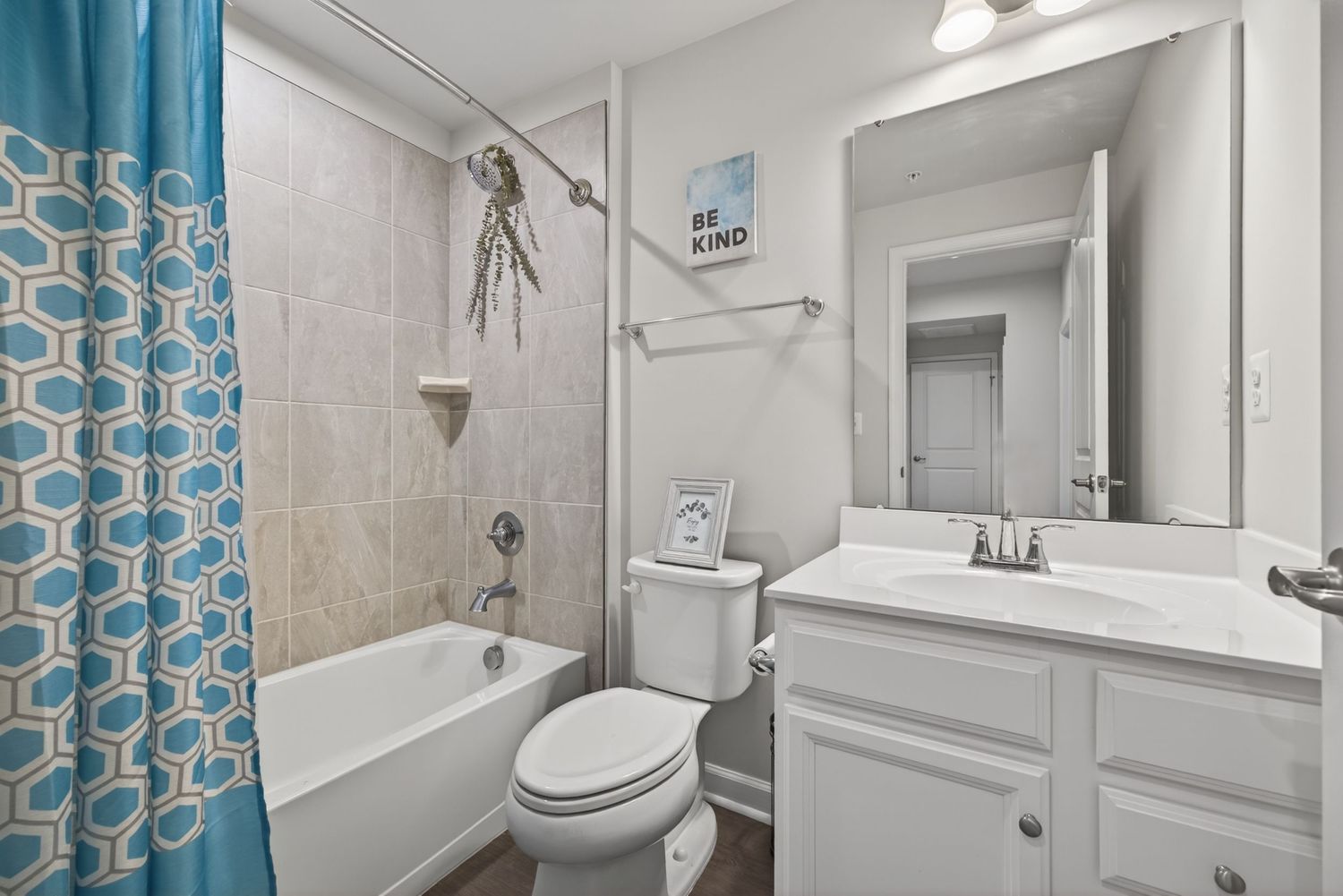 Bright bathroom with white vanity, large mirror, toilet, and tiled bathtub with blue geometric shower curtain.
