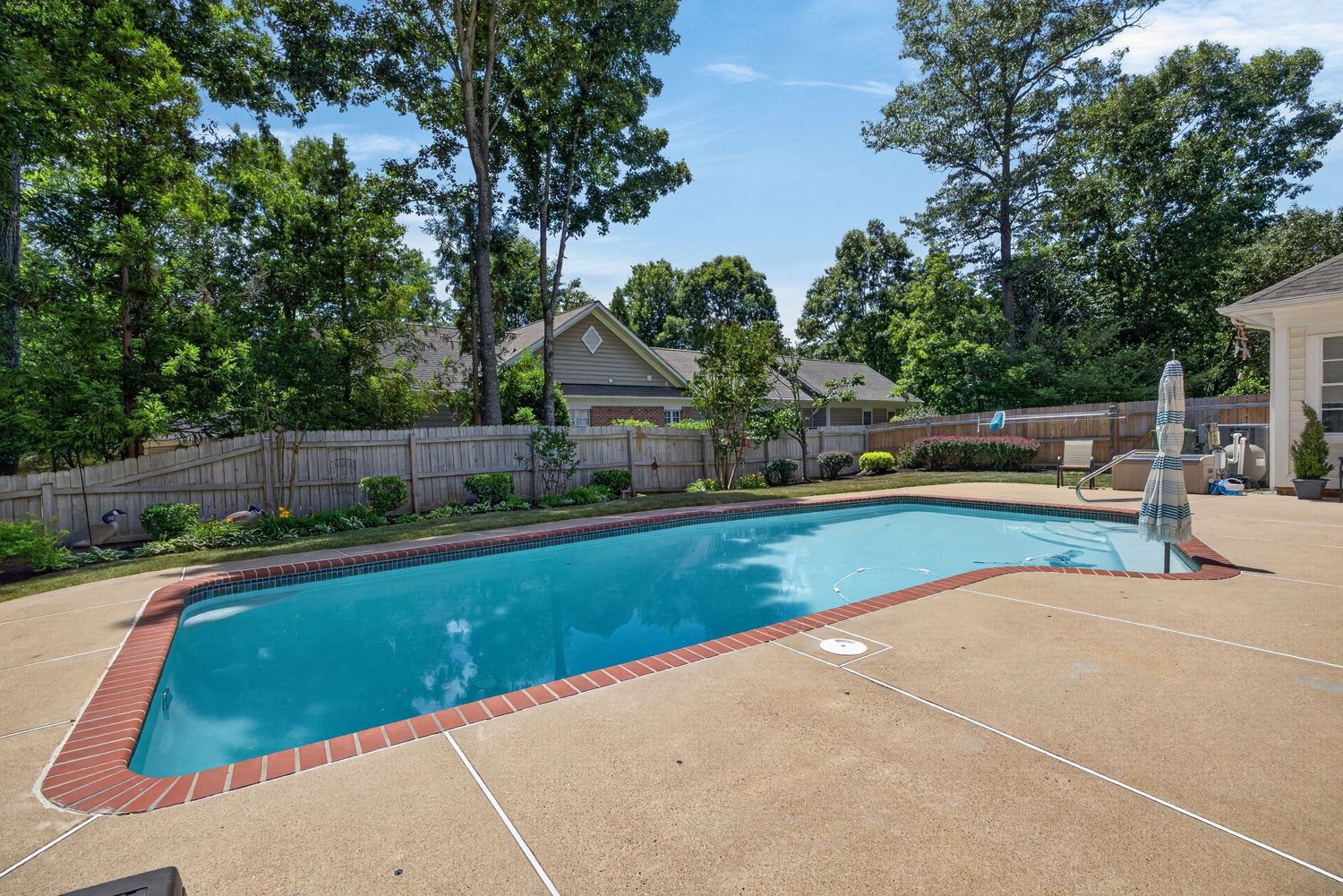 Outdoor rectangular swimming pool surrounded by brick edging and concrete patio with lush green trees and wooden fence backdrop.