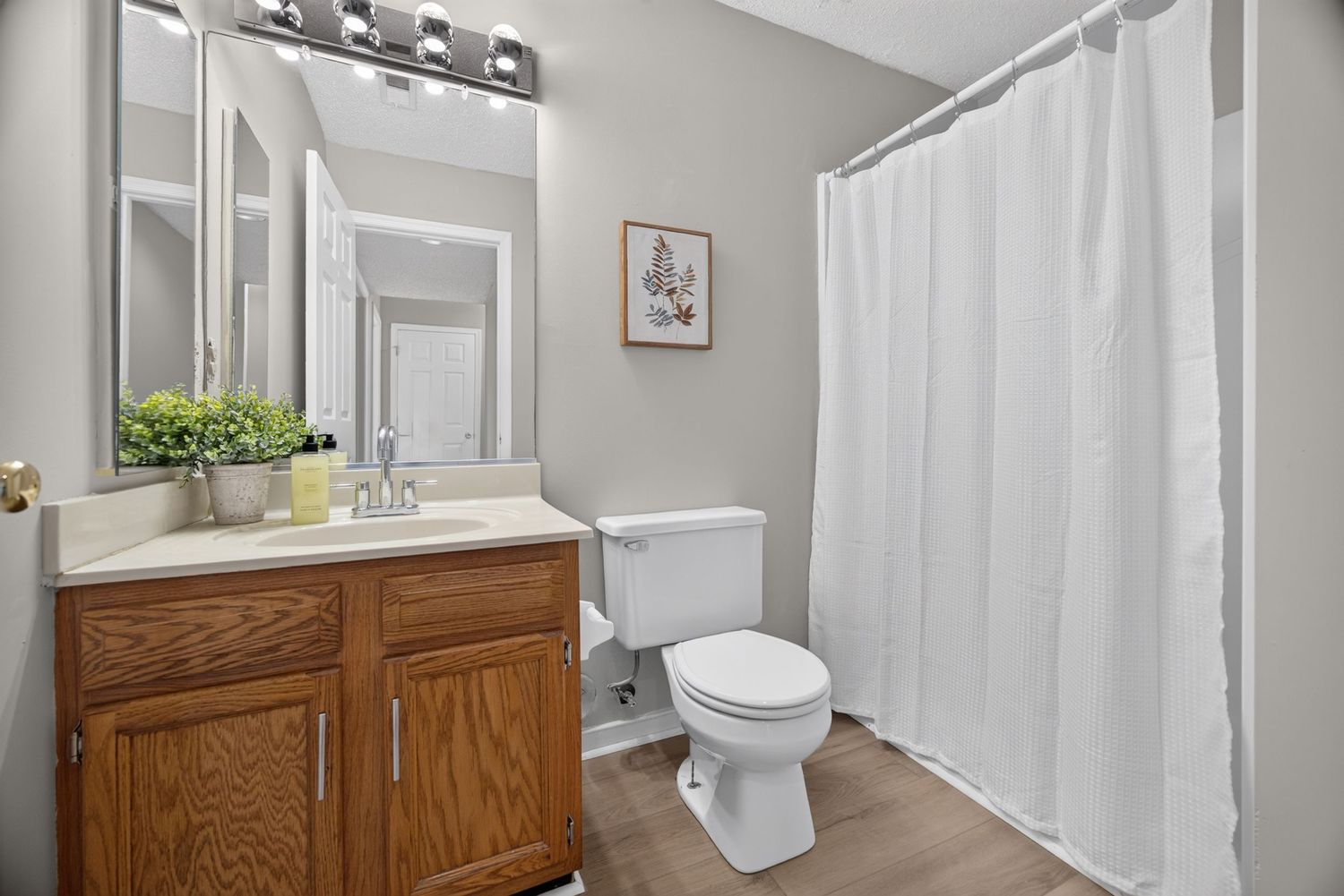 Clean bathroom with oak vanity, white toilet, shower with white curtain, and modern fixtures on light hardwood floors.
