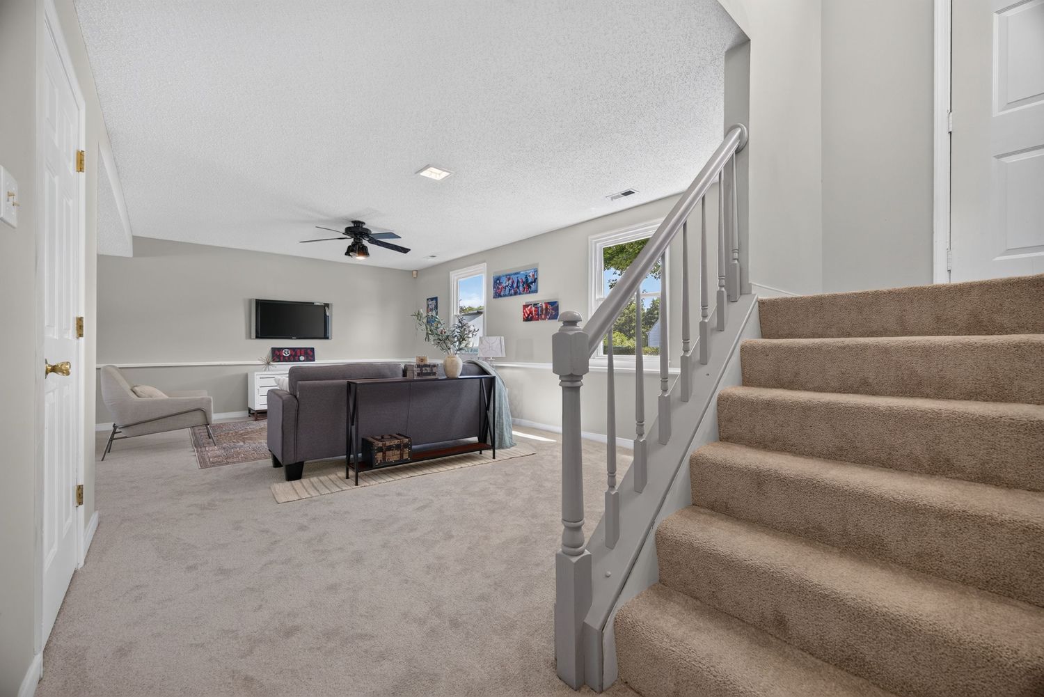 Bright living room with carpeted stairs, ceiling fan, gray sofa, and large windows providing natural light.