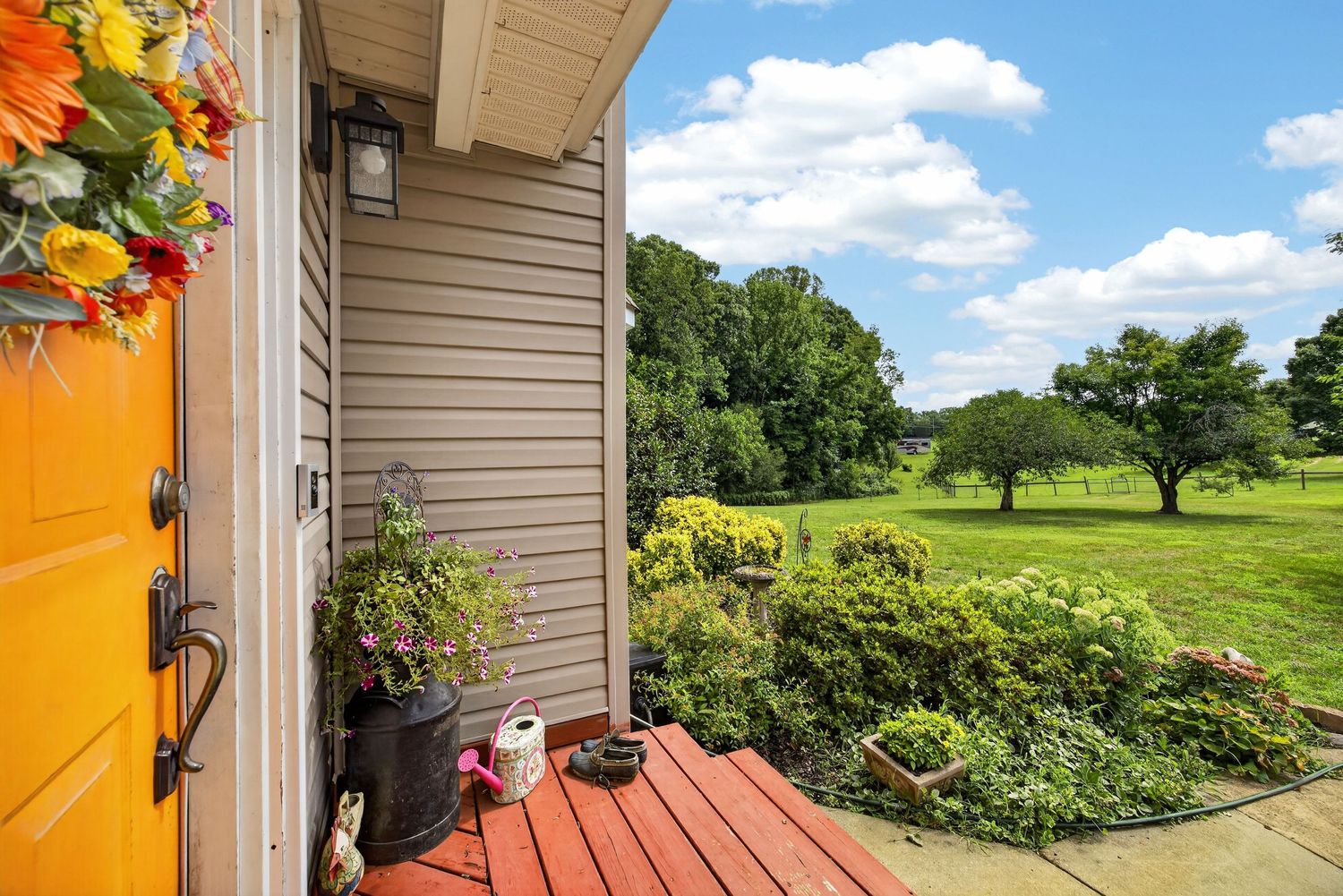 A charming home entryway featuring an orange front door, colorful wreath, potted plants, and a scenic green backyard view.