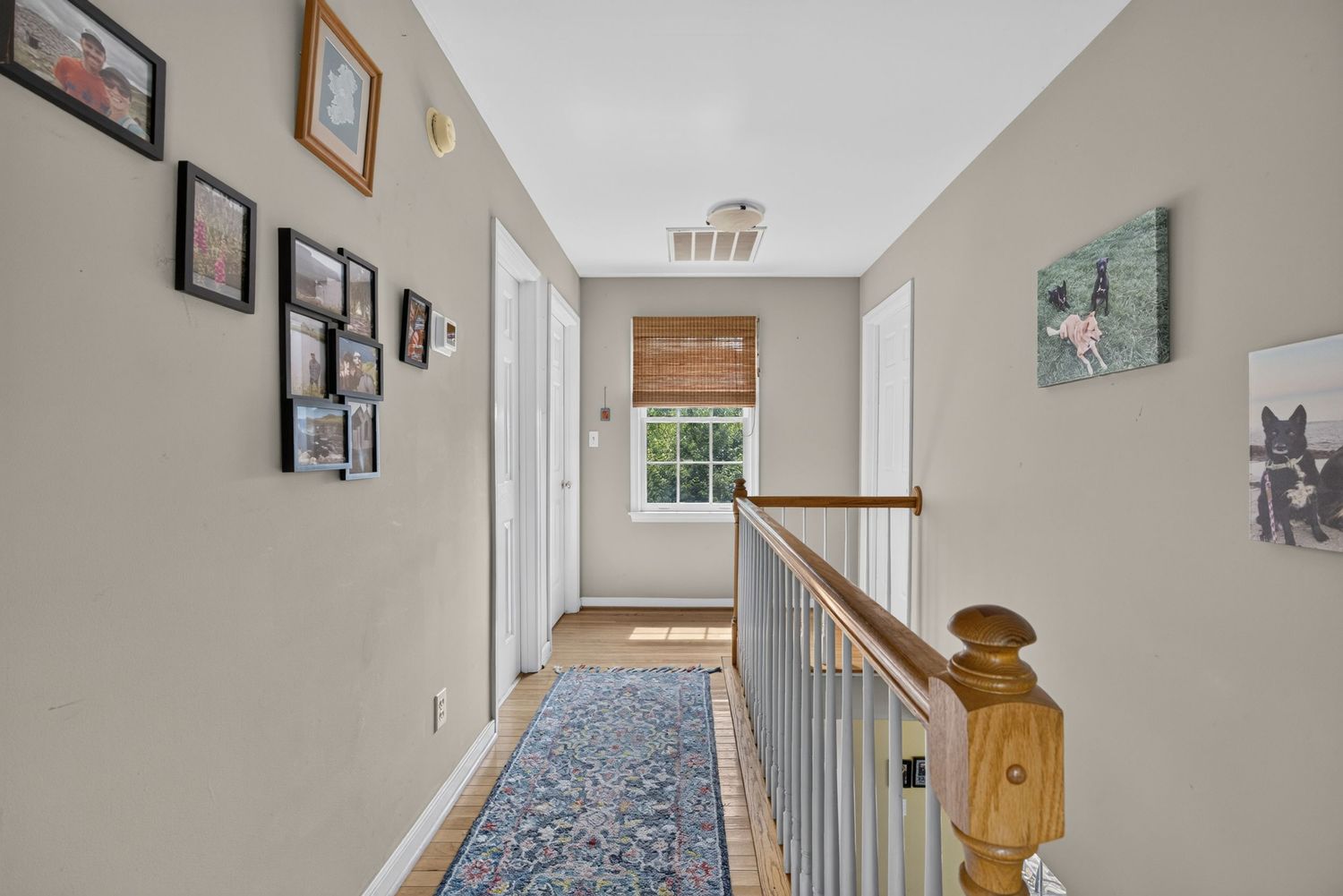 Bright upstairs hallway with wood railing, patterned runner rug, framed wall art, and natural light from a window at the end.