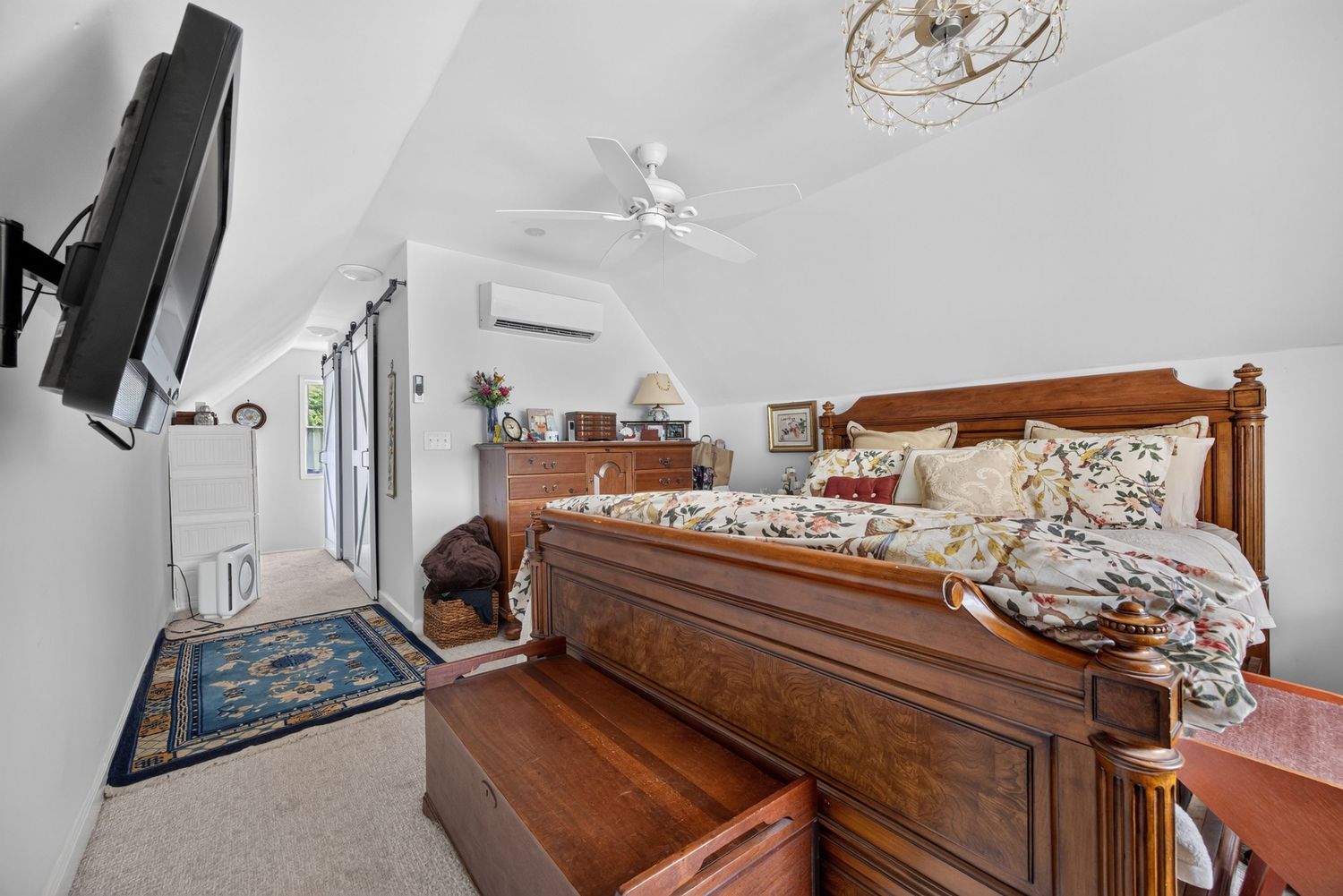 Spacious bedroom with vaulted ceiling, ceiling fan, wall-mounted TV, mini-split AC, and elegant wooden bed frame with floral bedding.