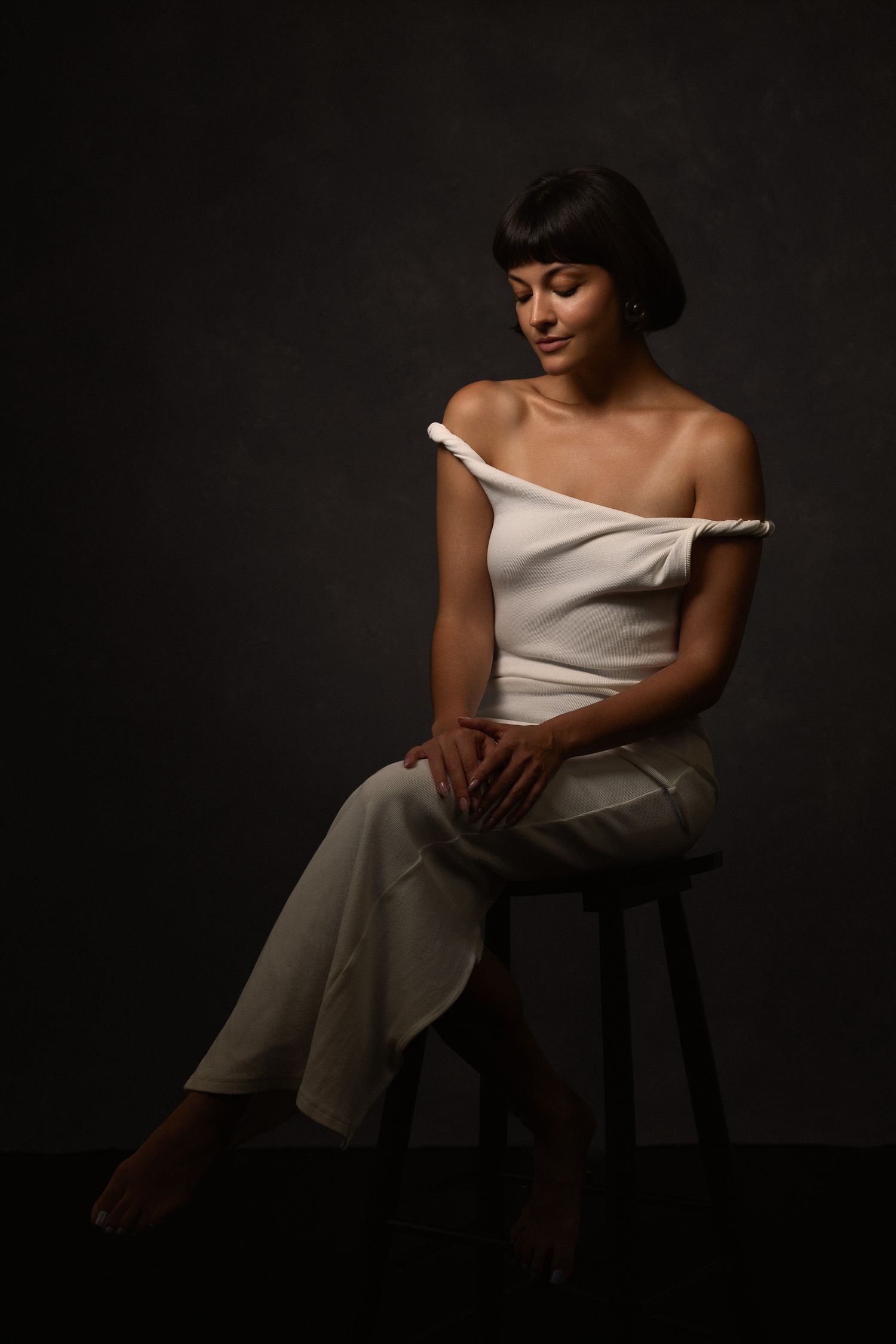 Elegant portrait in white off-shoulder top seated on dark stool.