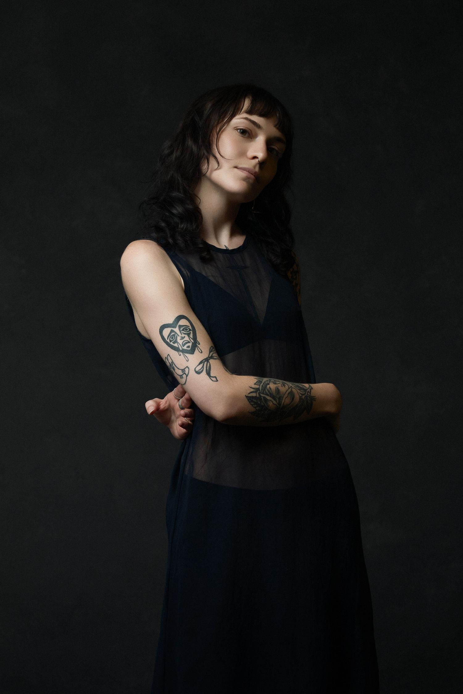 Artistic portrait of person in sheer black dress showing arm tattoos against dark background.