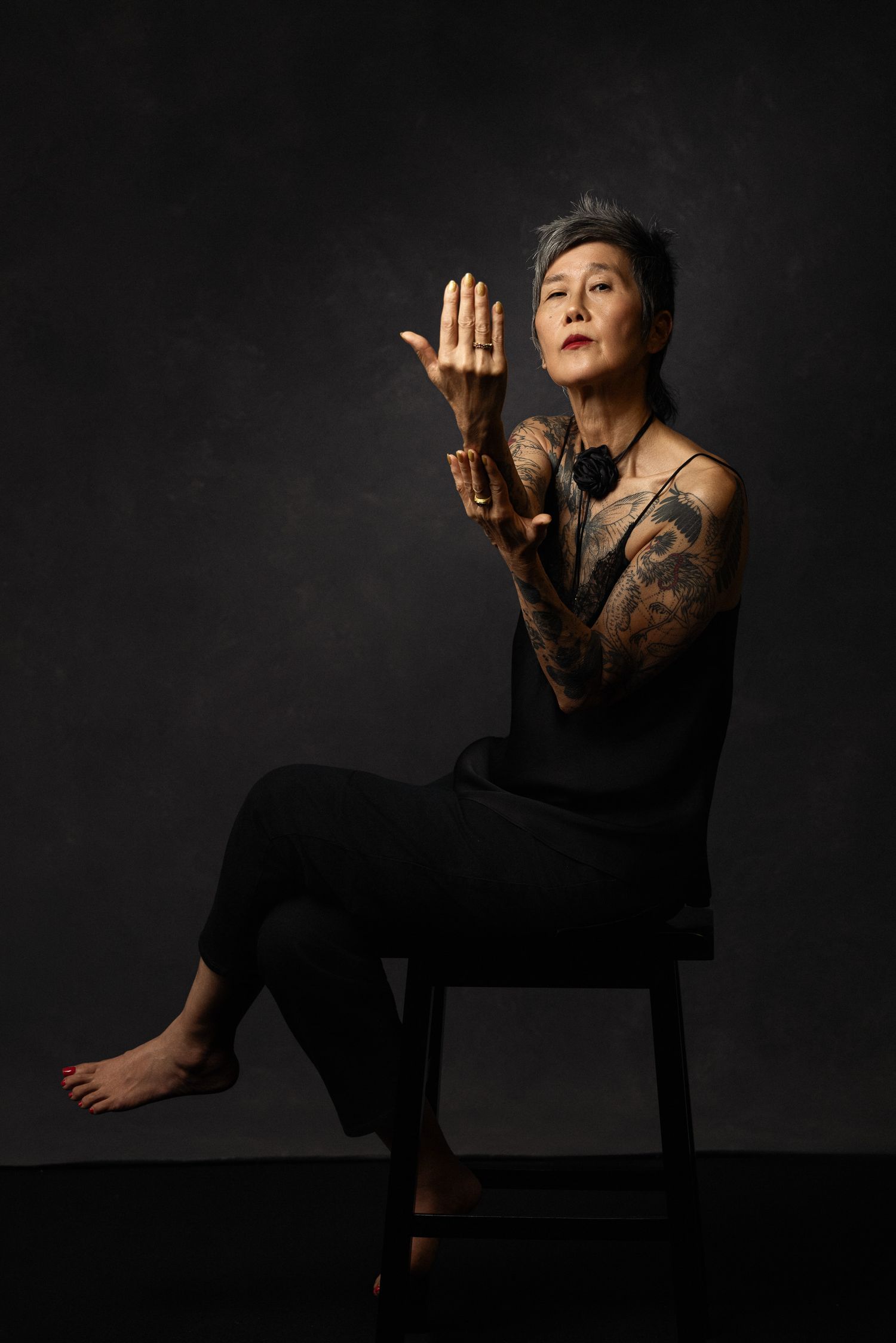 Artistic portrait of tattooed person in black clothing seated on chair against dark background.
