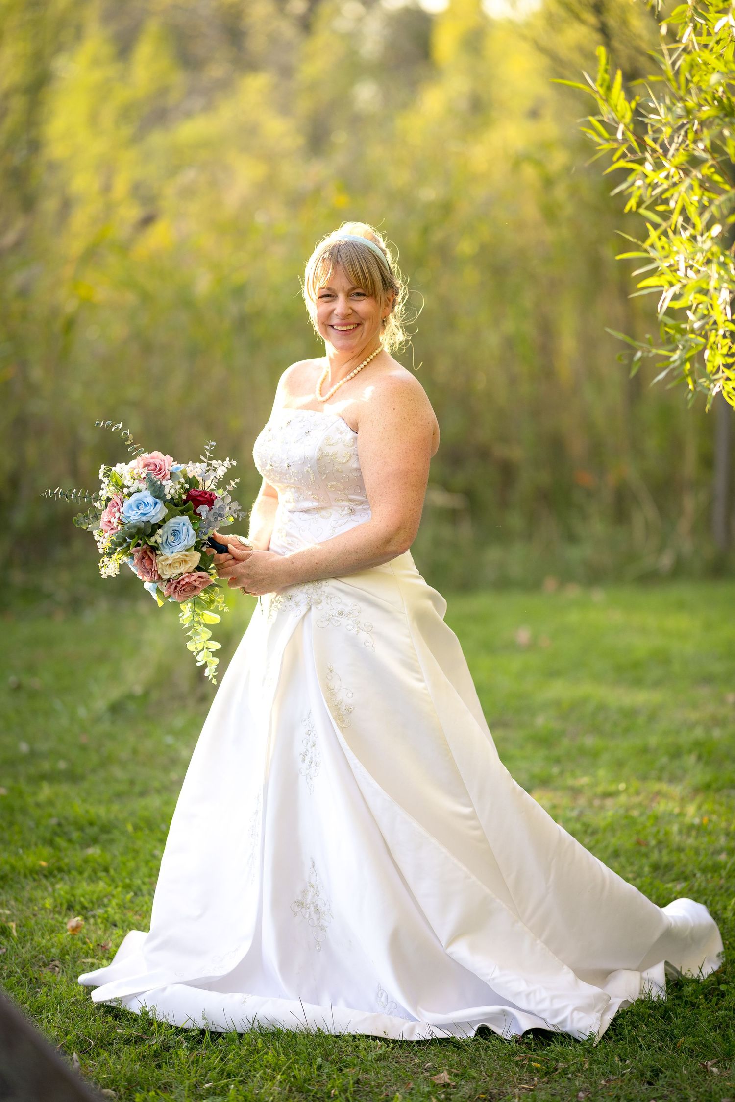 A bride in a white wedding dress stands gracefully on green grass holding a floral bouquet in a sunlit garden. Ottawa Wedding Photographer