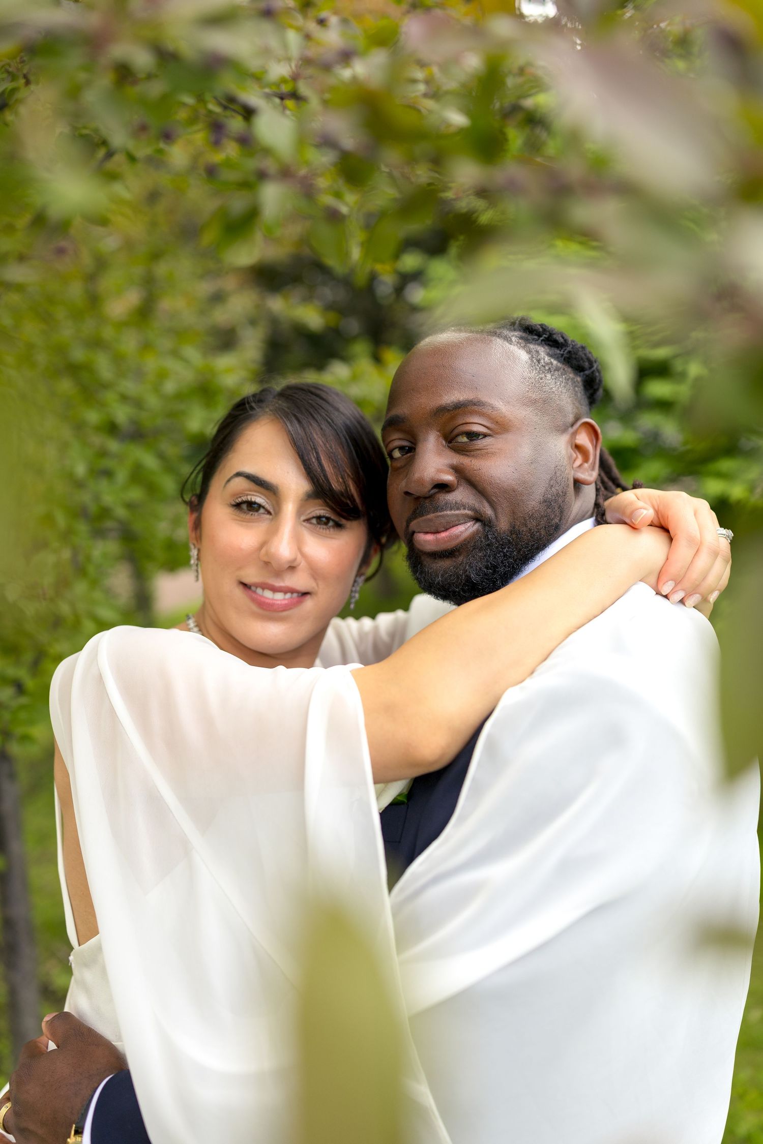 A couple in white wedding attire embracing outdoors, smiling against a lush green garden background. Ottawa Wedding Photographer