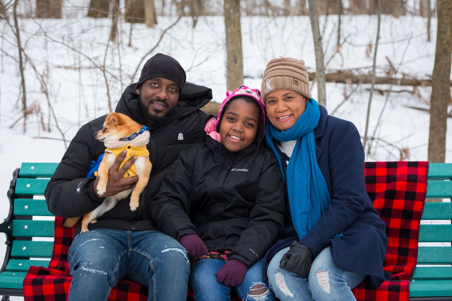 Family sitting on a snowy park bench with their small dog, bundled up in winter clothing.