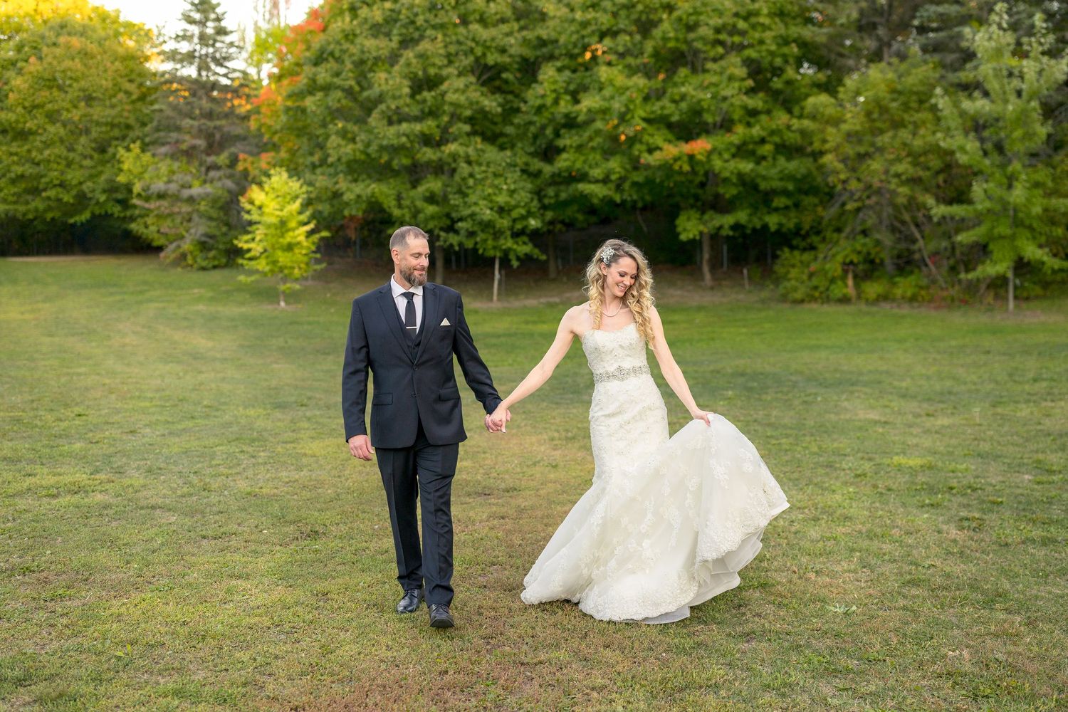 A bride & groom walks hand in hand across a green lawn with lush trees in the background on their wedding day. Ottawa Wedding Photography