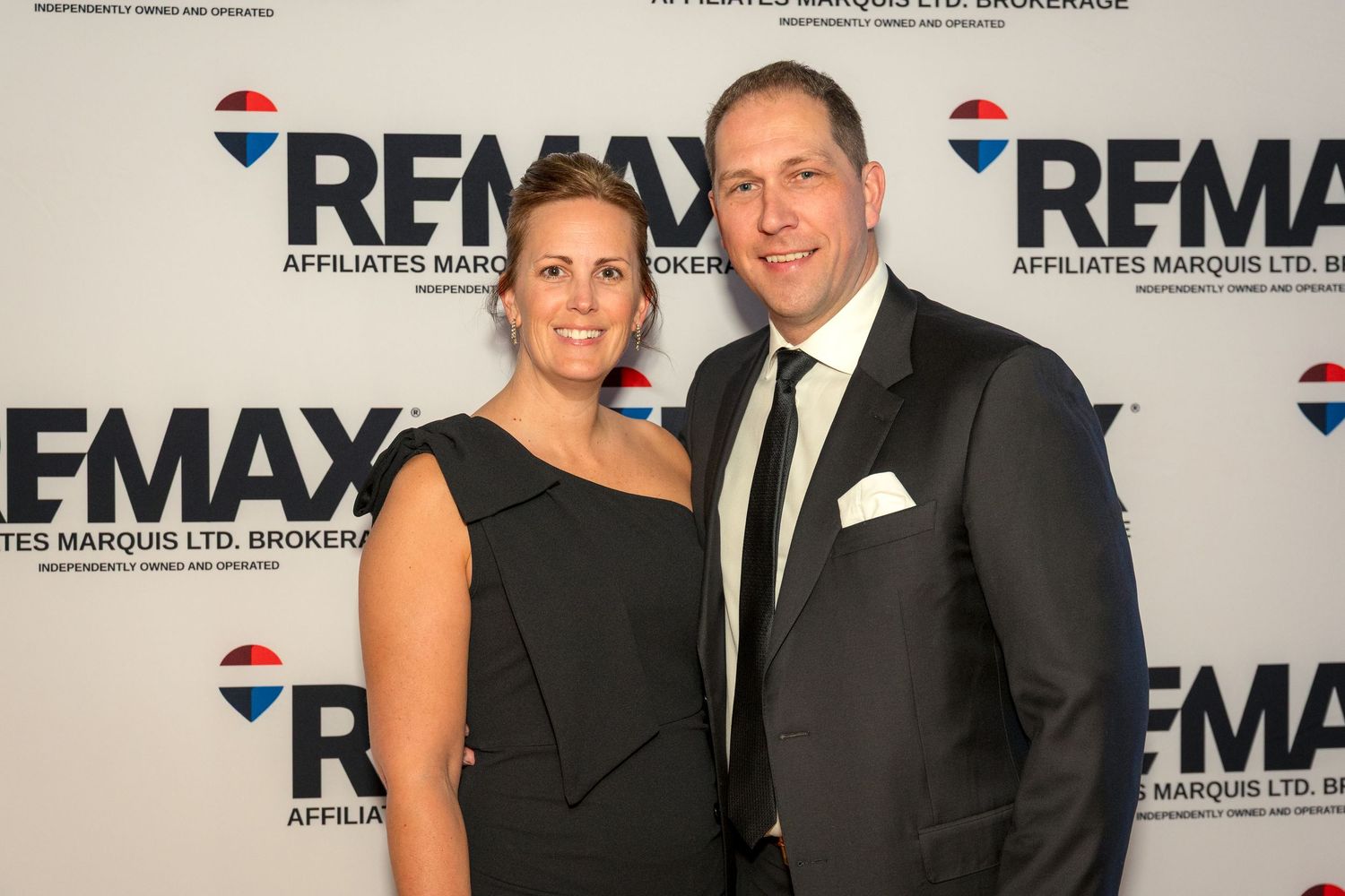 Real estate professionals in formal black attire pose together at a REMAX corporate event backdrop. Ottawa Event Photographer