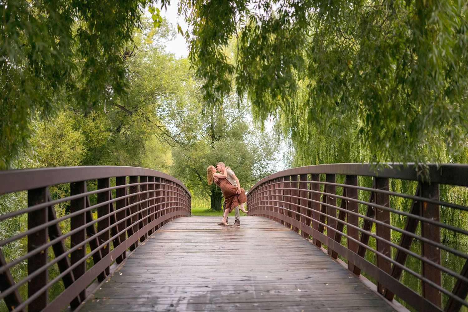 A couple shares a romantic moment on a wooden bridge surrounded by hanging willow trees. Ottawa Wedding Photographer