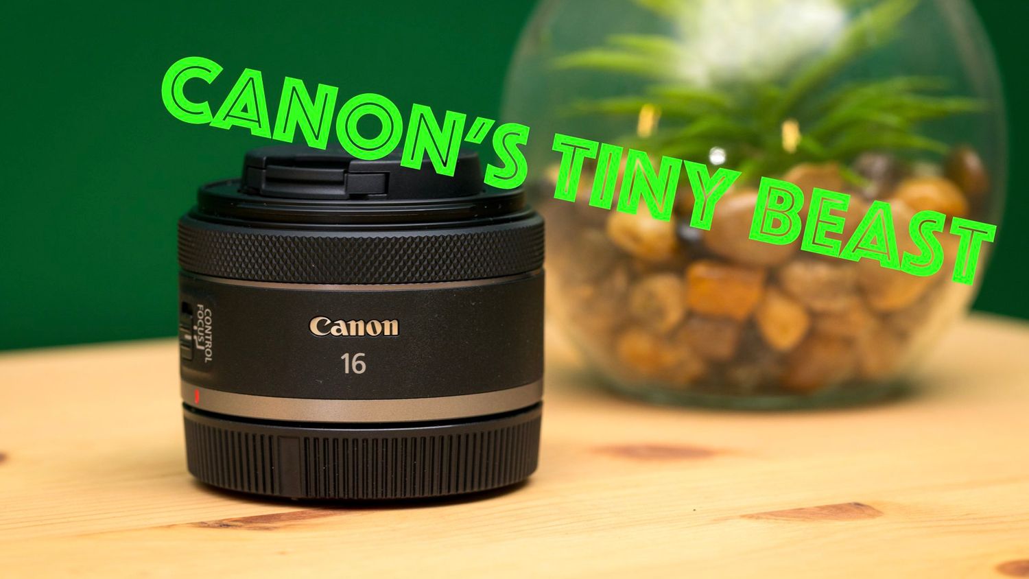 A Canon 16mm prime lens with green neon text effects displayed on a wooden table with a blurred glass bowl background.