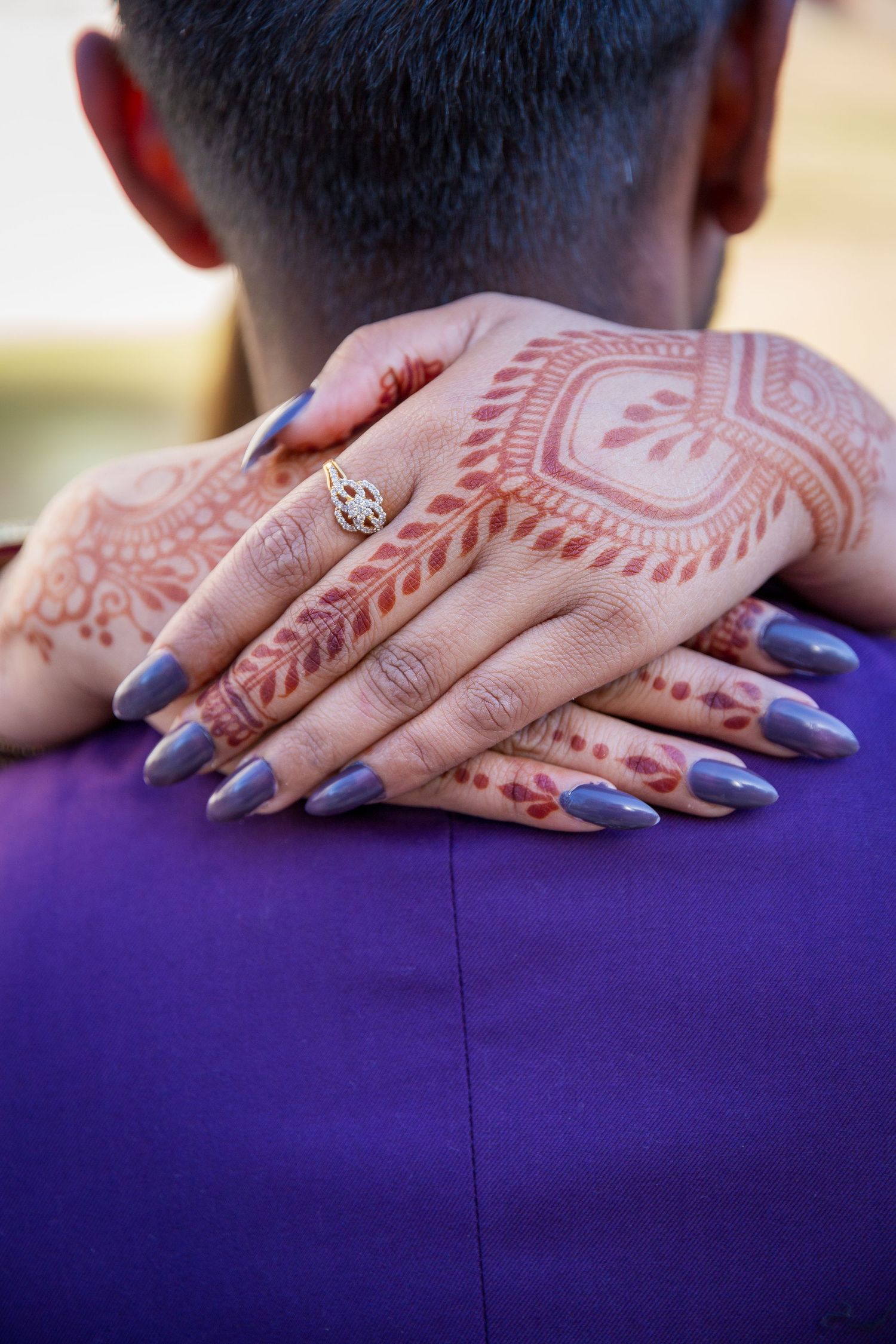 A close-up of hands with intricate henna design and purple nail polish embracing someone in a purple jacket.