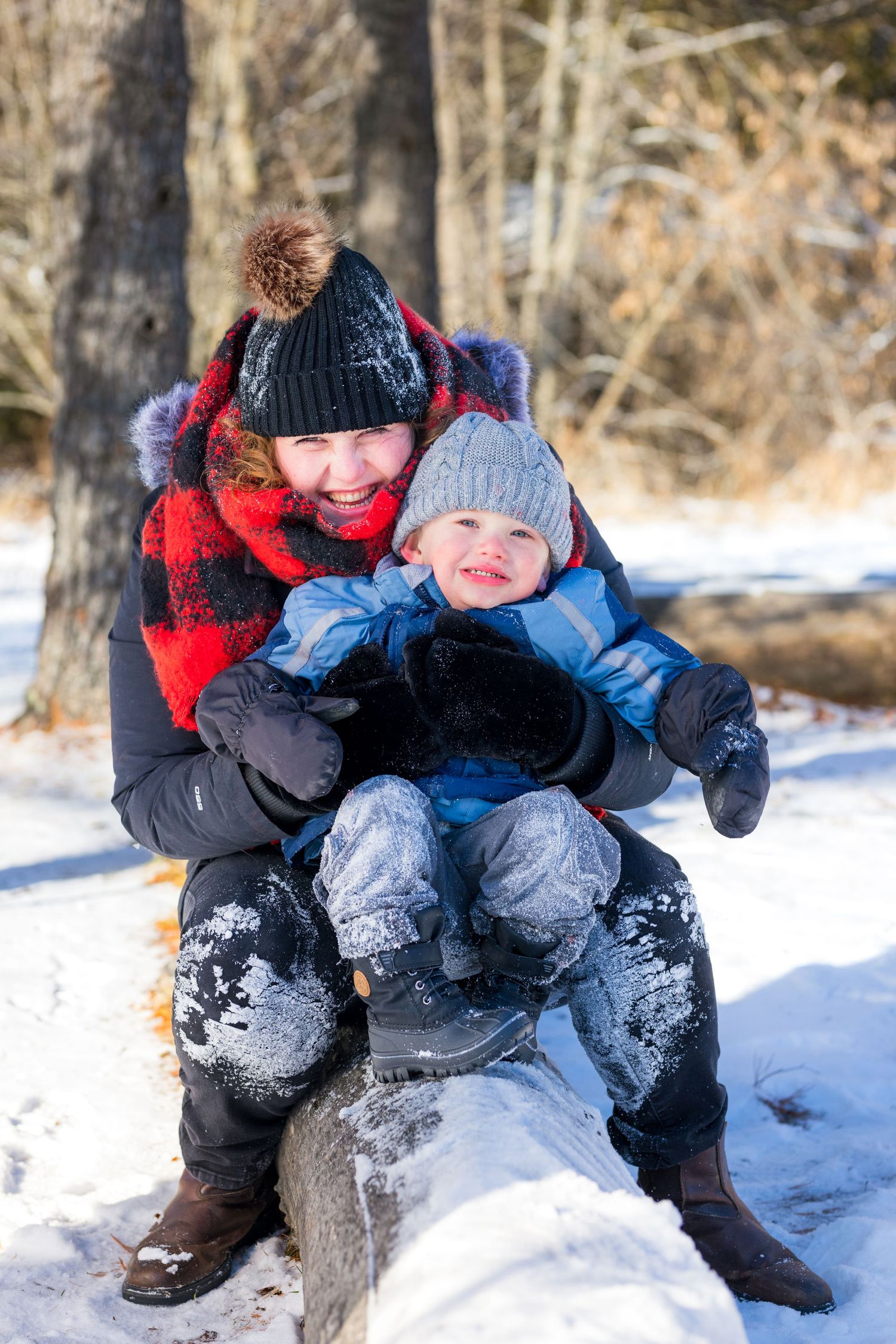 Two people sitting together on a log in winter snow wearing warm coats and winter hats while enjoying the outdoors.