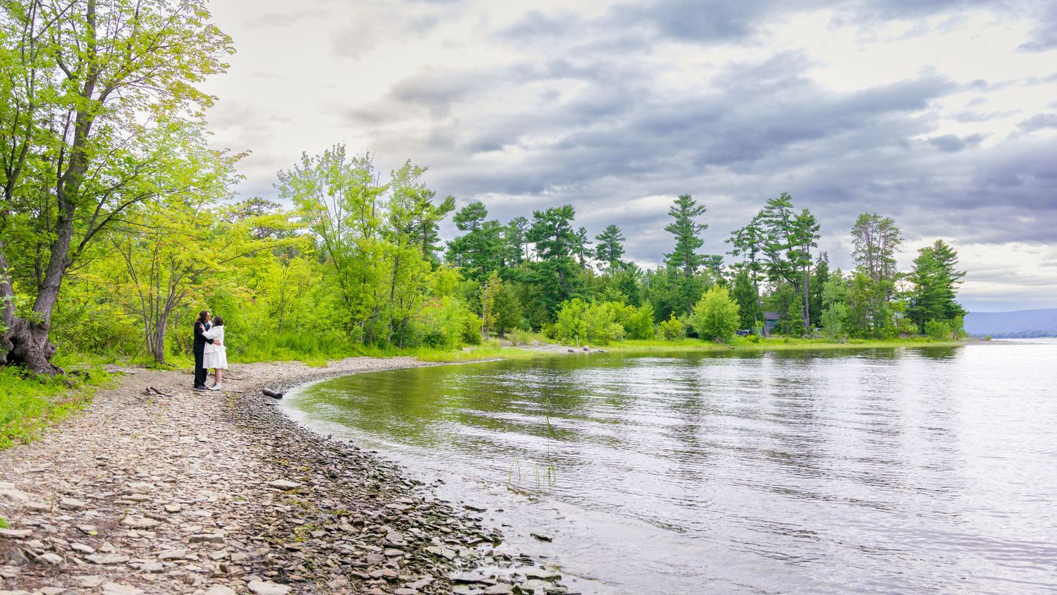 A scenic lakeside view with figures walking along a rocky shore under cloudy skies. Ottawa Wedding Photographer