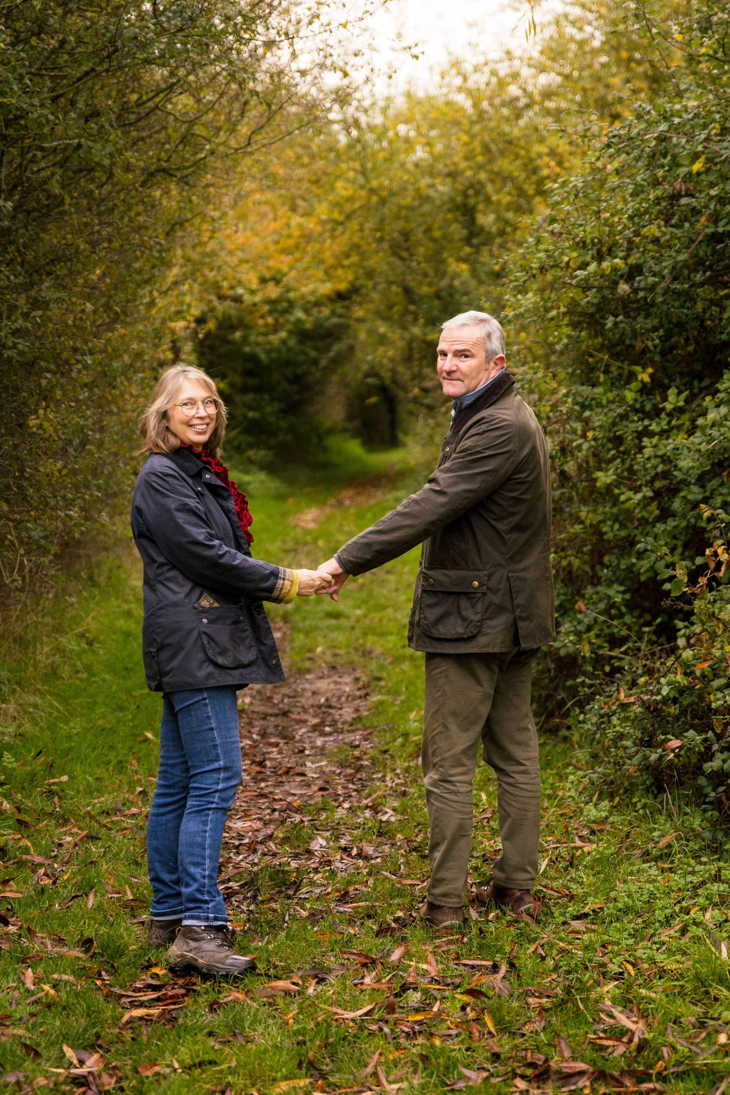 Couple holding hands on autumn path surrounded by golden leaves and green hedges.