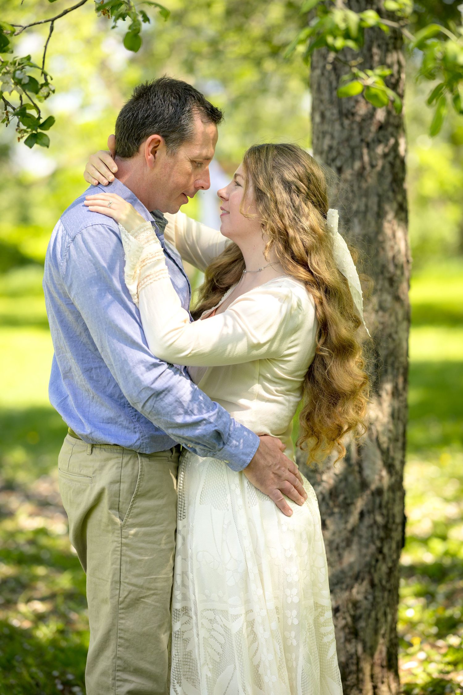 Couple in romantic embrace outdoors, bride in white gown with long hair, groom in blue shirt, surrounded by lush green trees. Ottawa Family